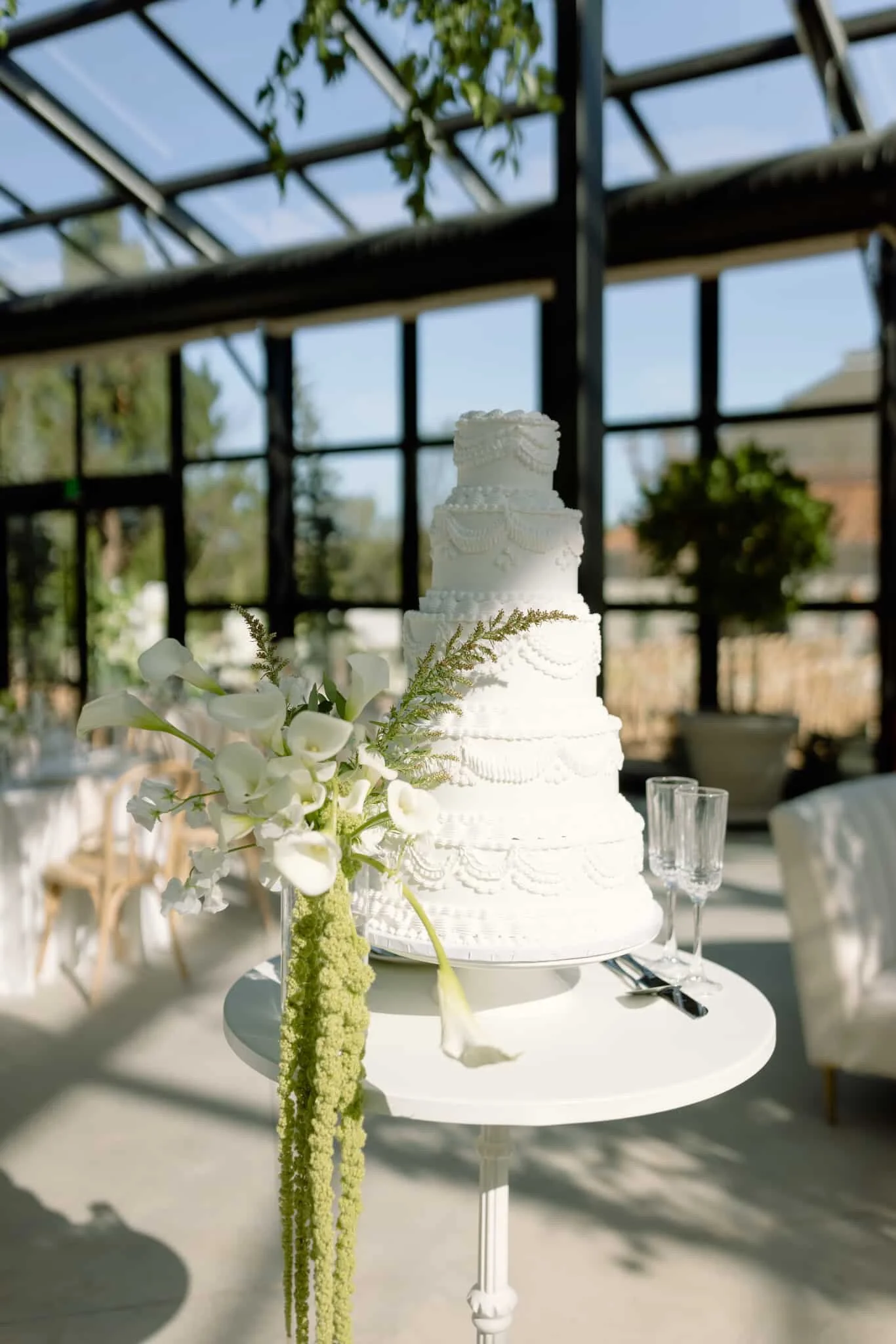A large white wedding cake decorated with intricate icing patterns and beaded accents, sitting on a white round table with a floral arrangement of white calla lilies and greenery in a glass vase. The background shows a glass-walled venue with a view 