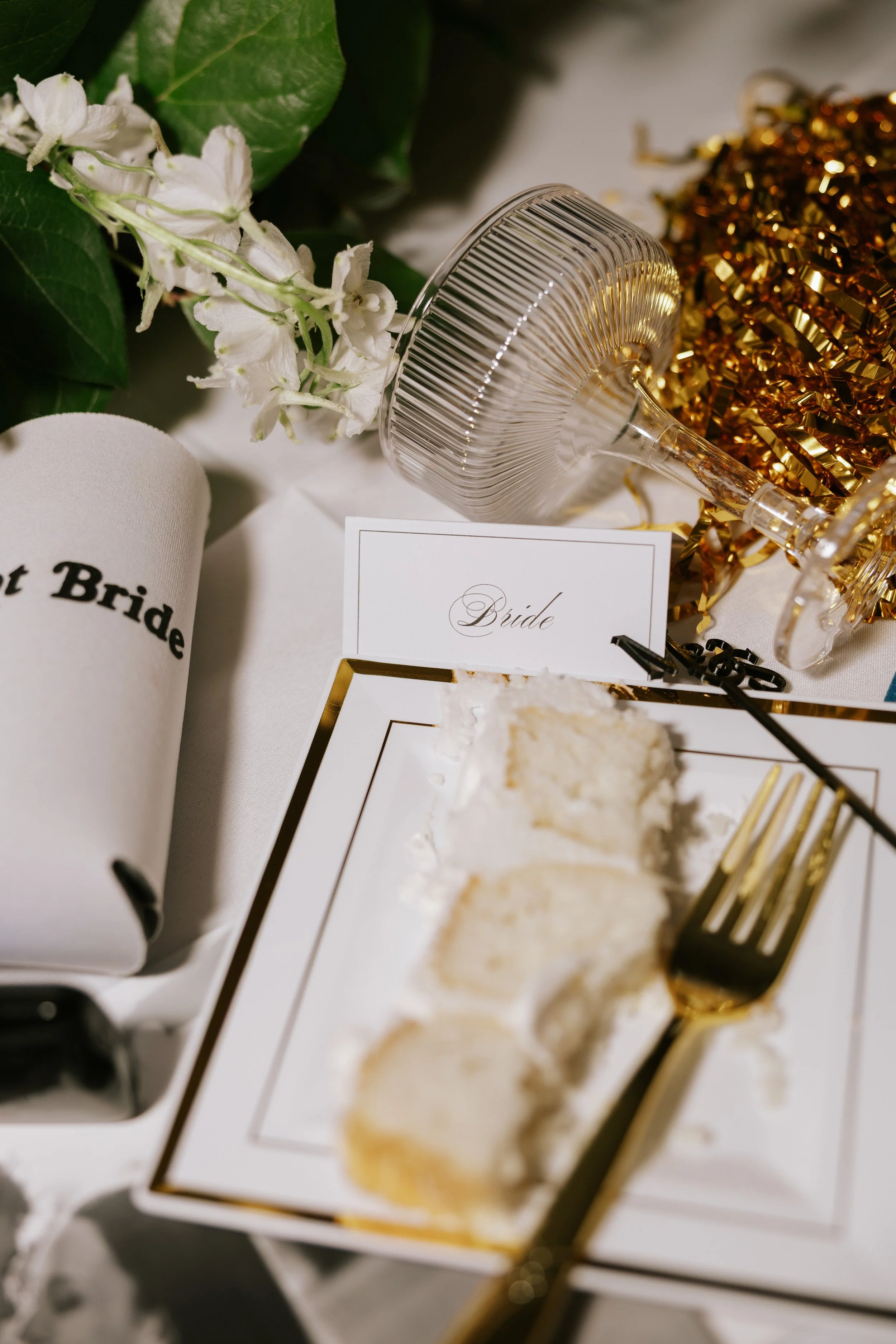 A wedding reception table setting with a slice of cake, a wedding fork, a champagne glass, a 'Bride' card, decorative golden and clear elements, and a napkin that says 'Not Bride', all arranged for a bride or bridal party celebration.