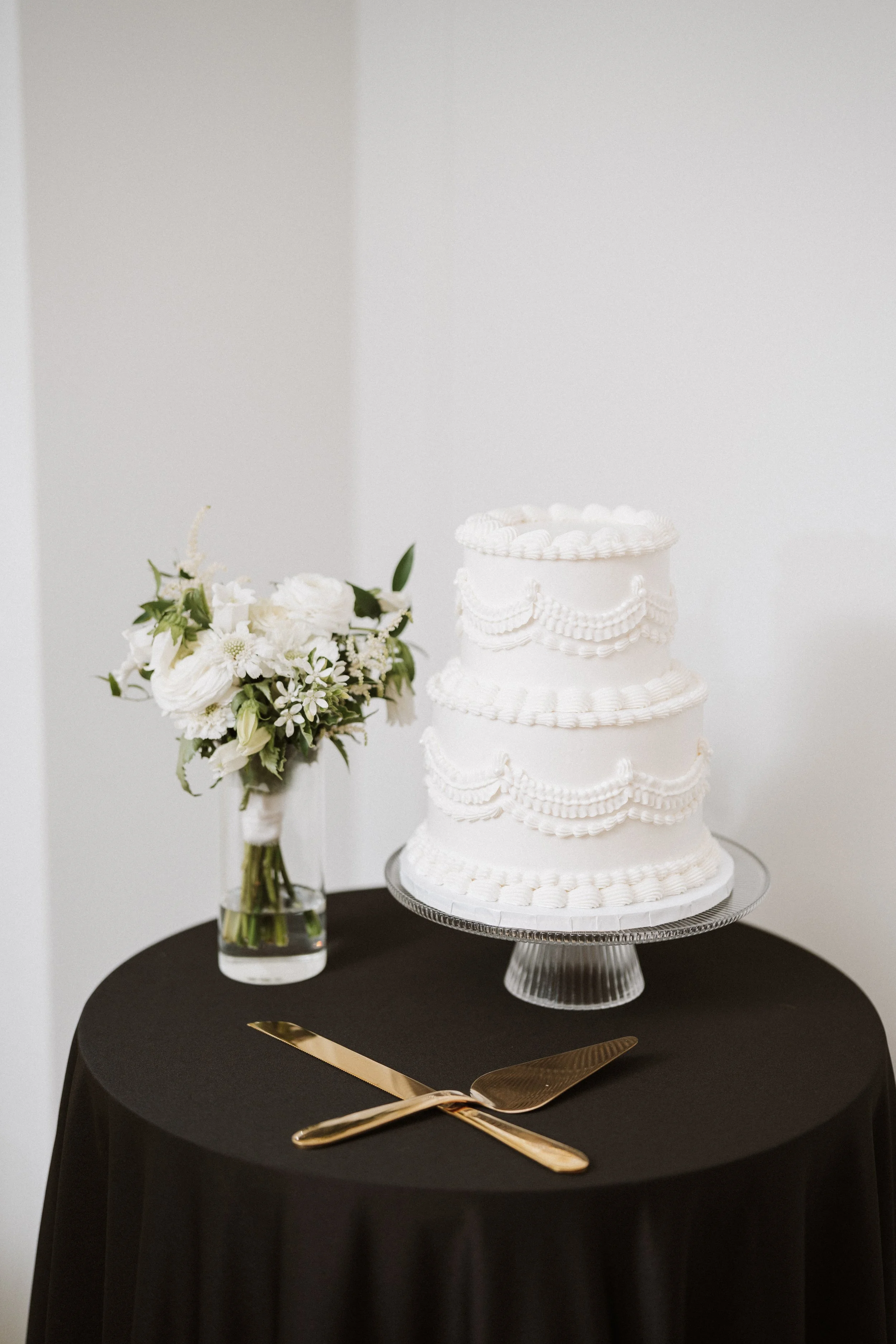 A three-tier white wedding cake with decorative piping and icing, placed on a glass cake stand, set on a black table cover. Next to the cake, there's a vase with a bouquet of white flowers and greenery. In front of the cake, a gold cake knife and server rest crossed on the table.