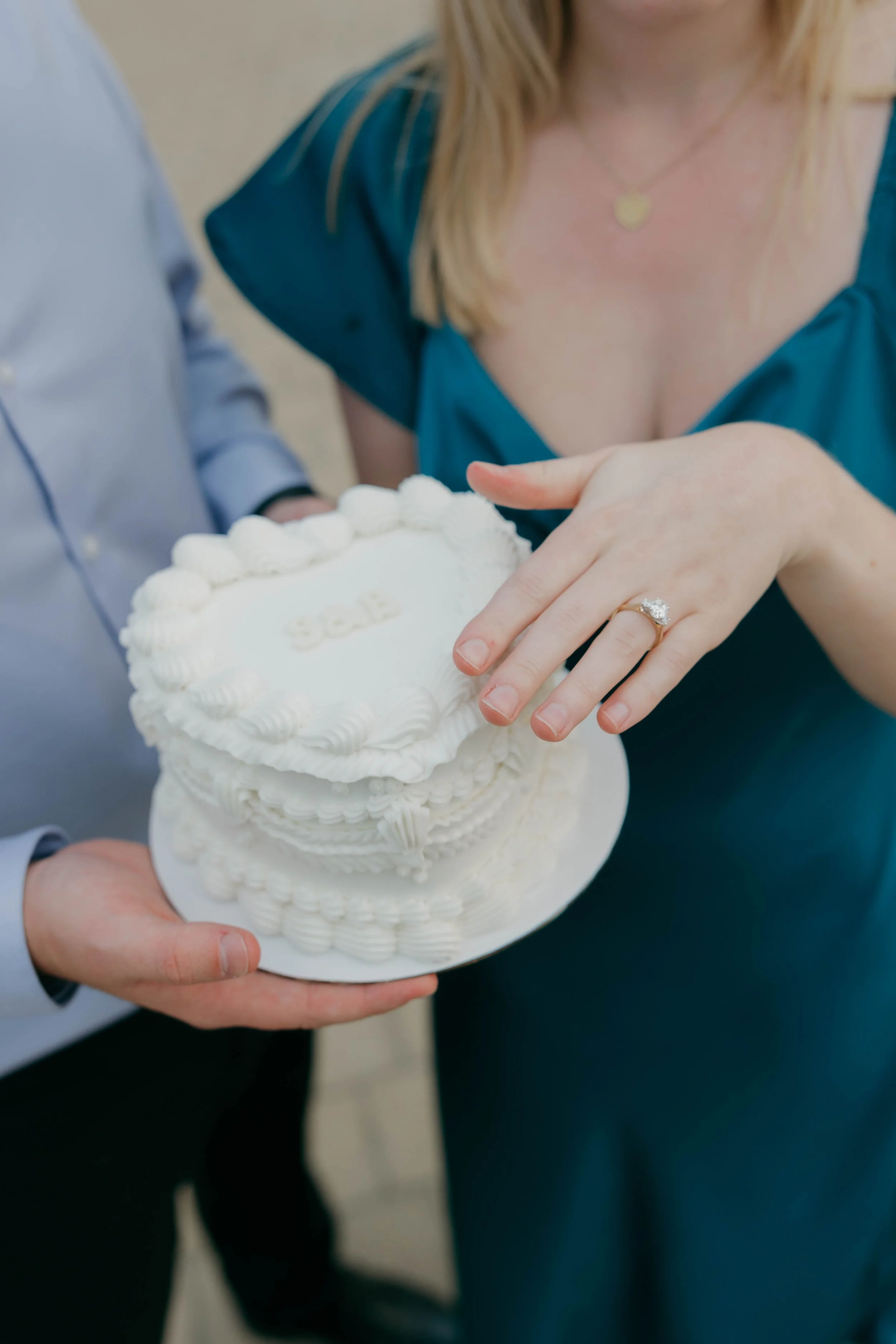 Close-up of a woman with an engagement ring on her finger, touching a heart-shaped white wedding cake with white icing decorations being held by a man.