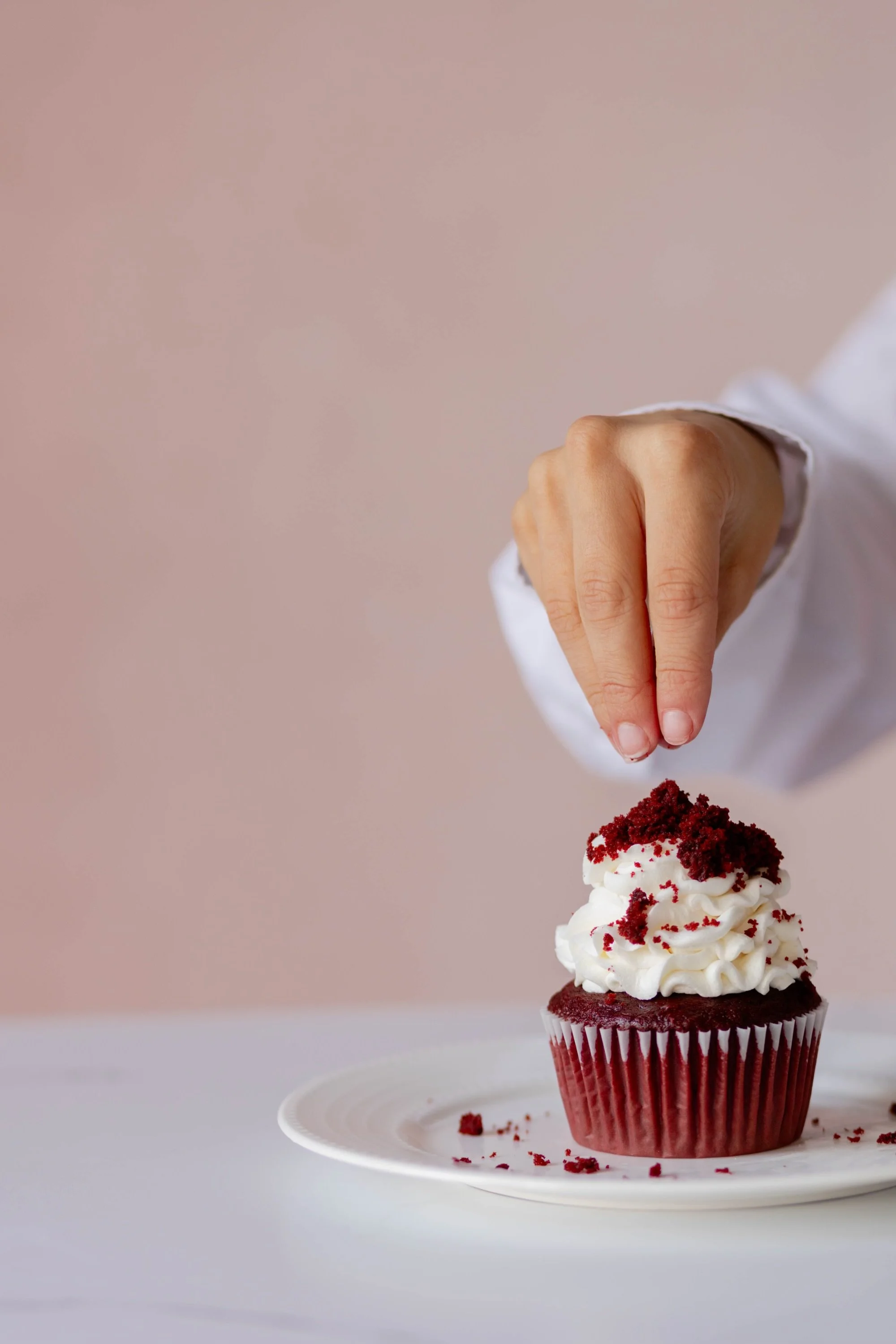 Person decorating a red velvet cupcake with whipped cream and red crumbs on a white plate.
