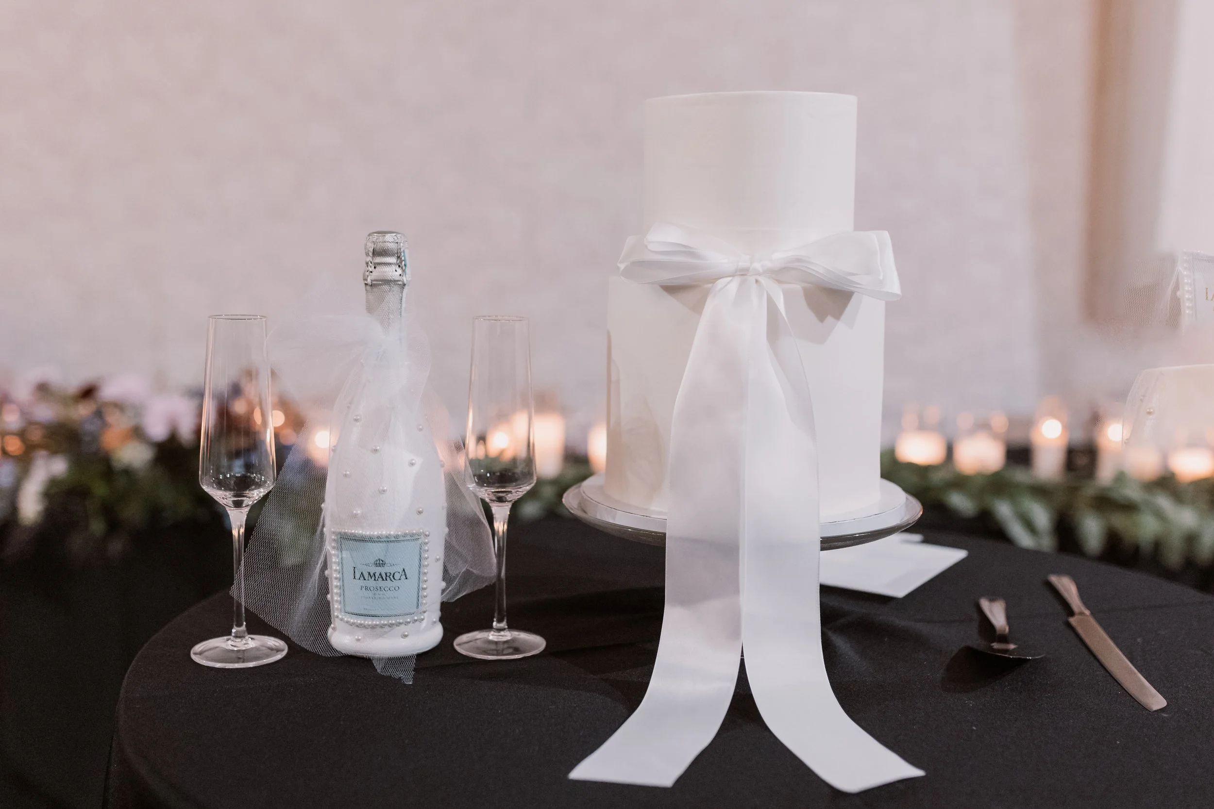 Wedding table setup with a white wedding cake decorated with a white ribbon, a bottle of Lamarka Prosecco wrapped in a tulle bag, two champagne flutes, a spoon, and a knife on a black tablecloth, with candles and floral decorations in the background.