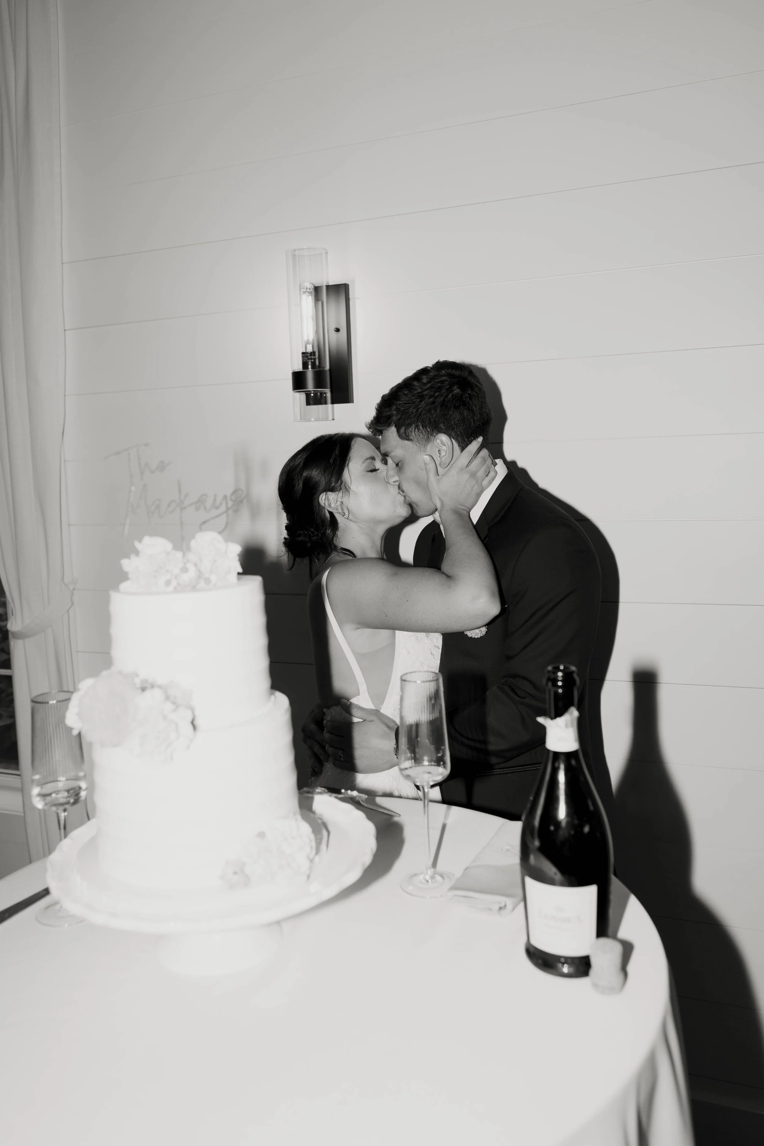 A bride and groom kiss at their wedding reception, with a wedding cake, champagne, and glasses on the table, in black and white.