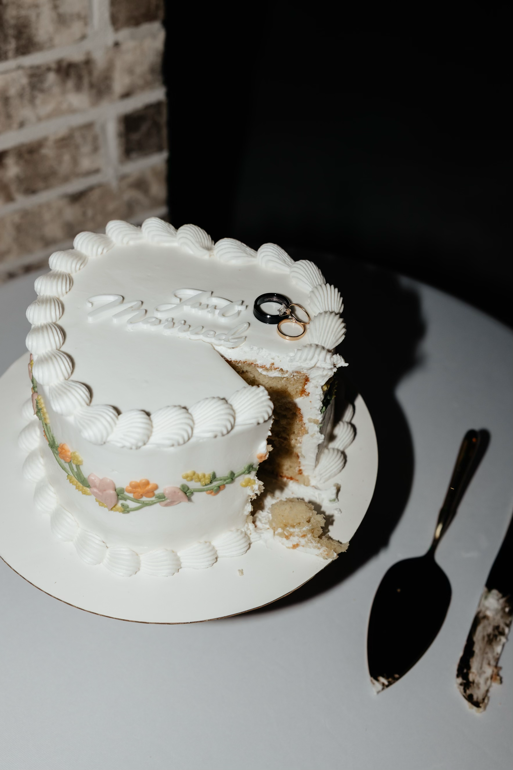 A white wedding cake with pastel-colored floral decorations, two rings on top, with a slice removed, placed on a cake stand on a table with a black cake server and knife nearby.