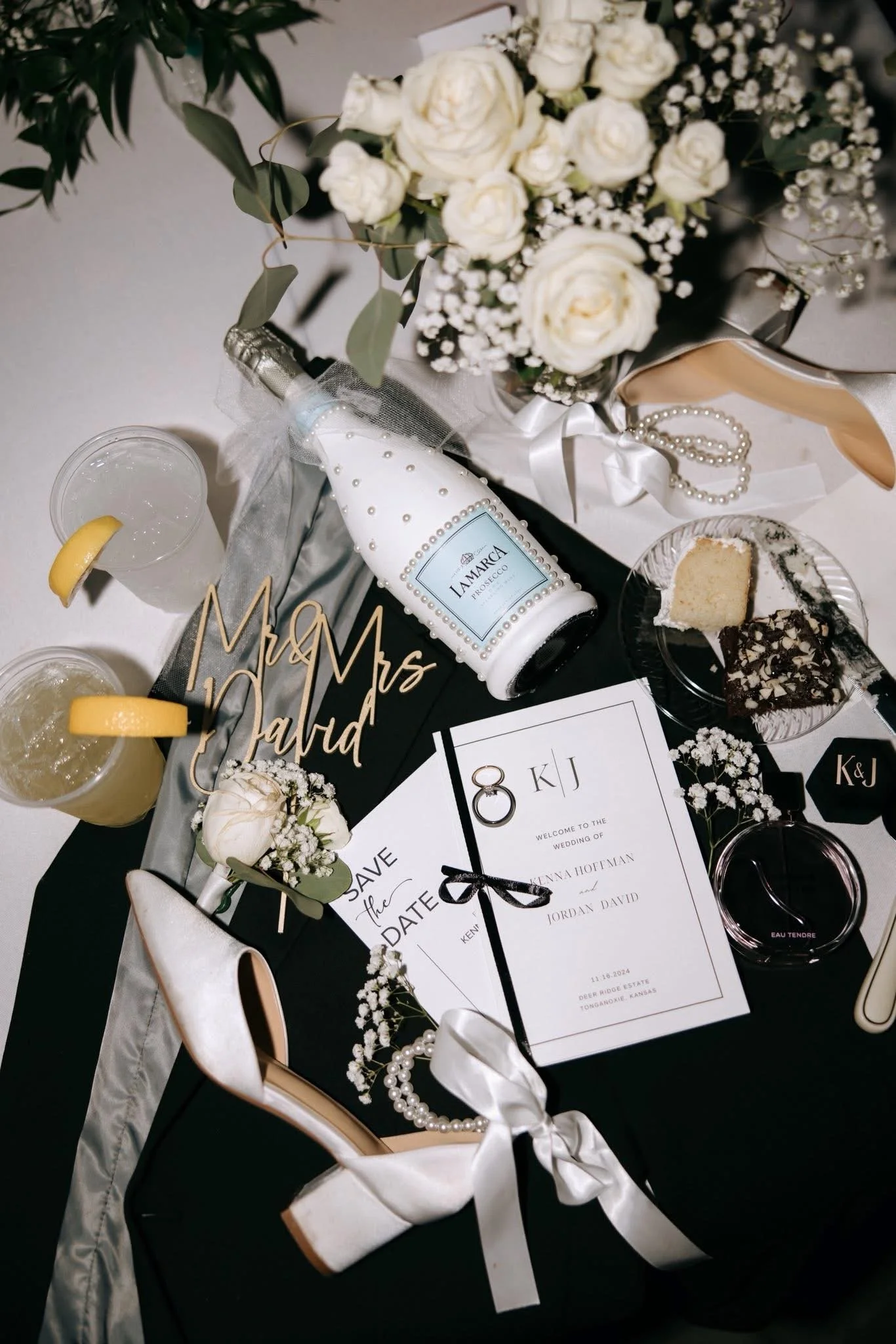 Wedding table decorations featuring a bouquet of white roses and baby's breath, a bottle of La Marca Prosecco with pearl decorations, wedding rings on the table, a 'Mr & Mrs' sign, a 'Save the Date' card, wedding candies, two glasses with lemon slice