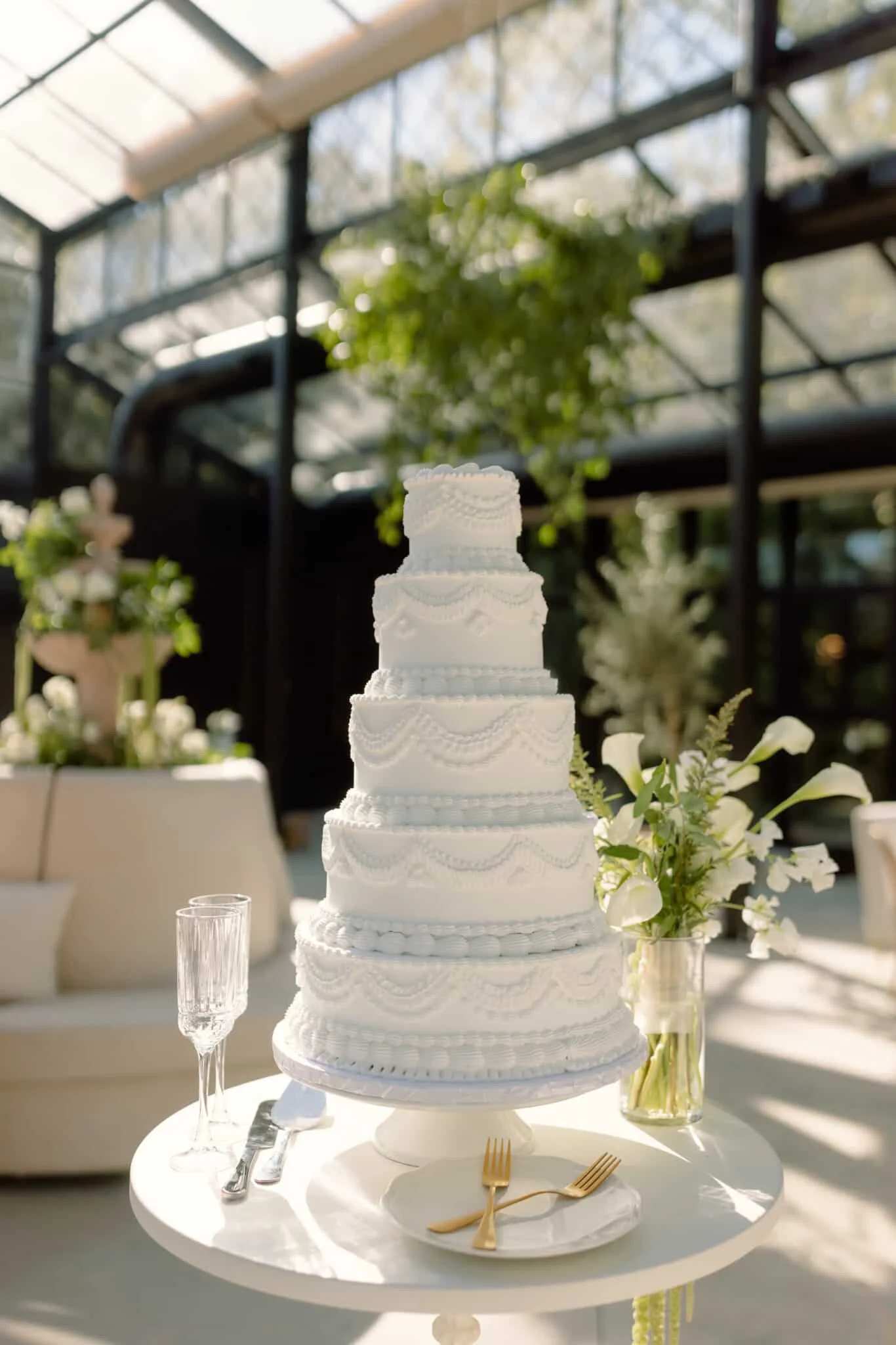 A tall, white, multi-tiered wedding cake with intricate piping decorations, placed on a white round table with gold forks, a marble cake stand, and glasses. A vase of white flowers is next to the cake, and the background features a glass greenhouse w