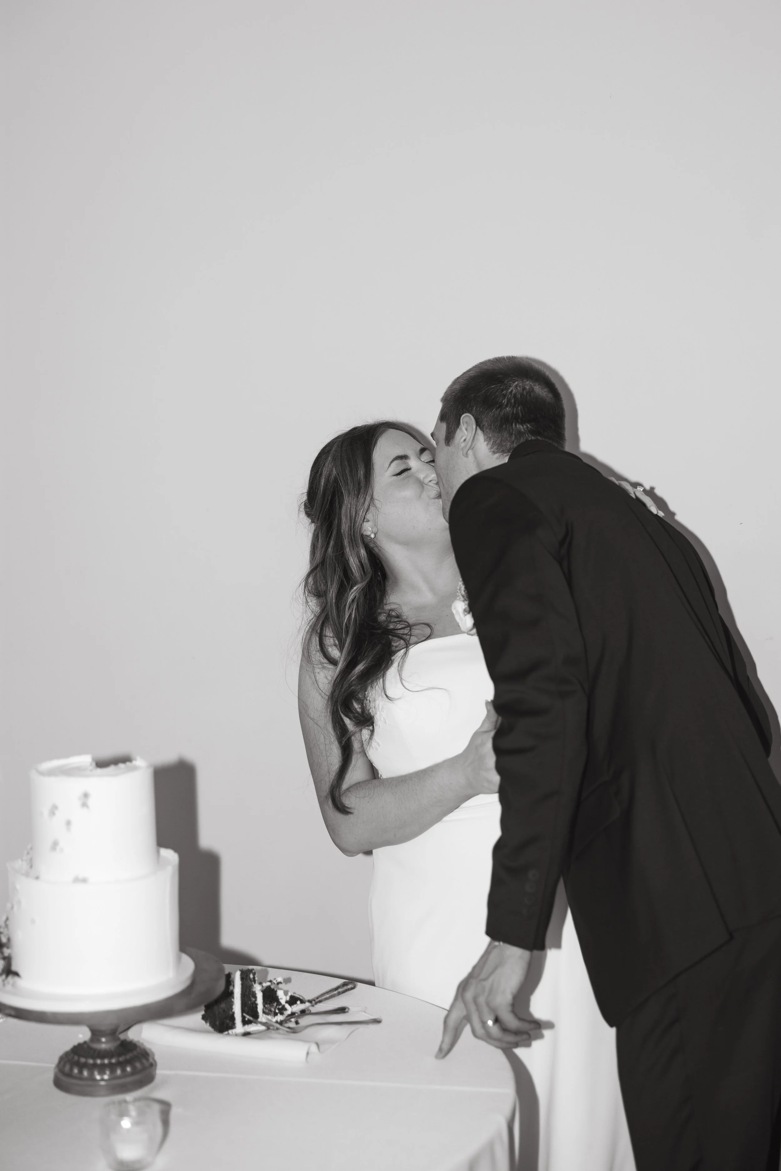 A couple sharing a kiss at their wedding reception, with a wedding cake on the table in front of them.