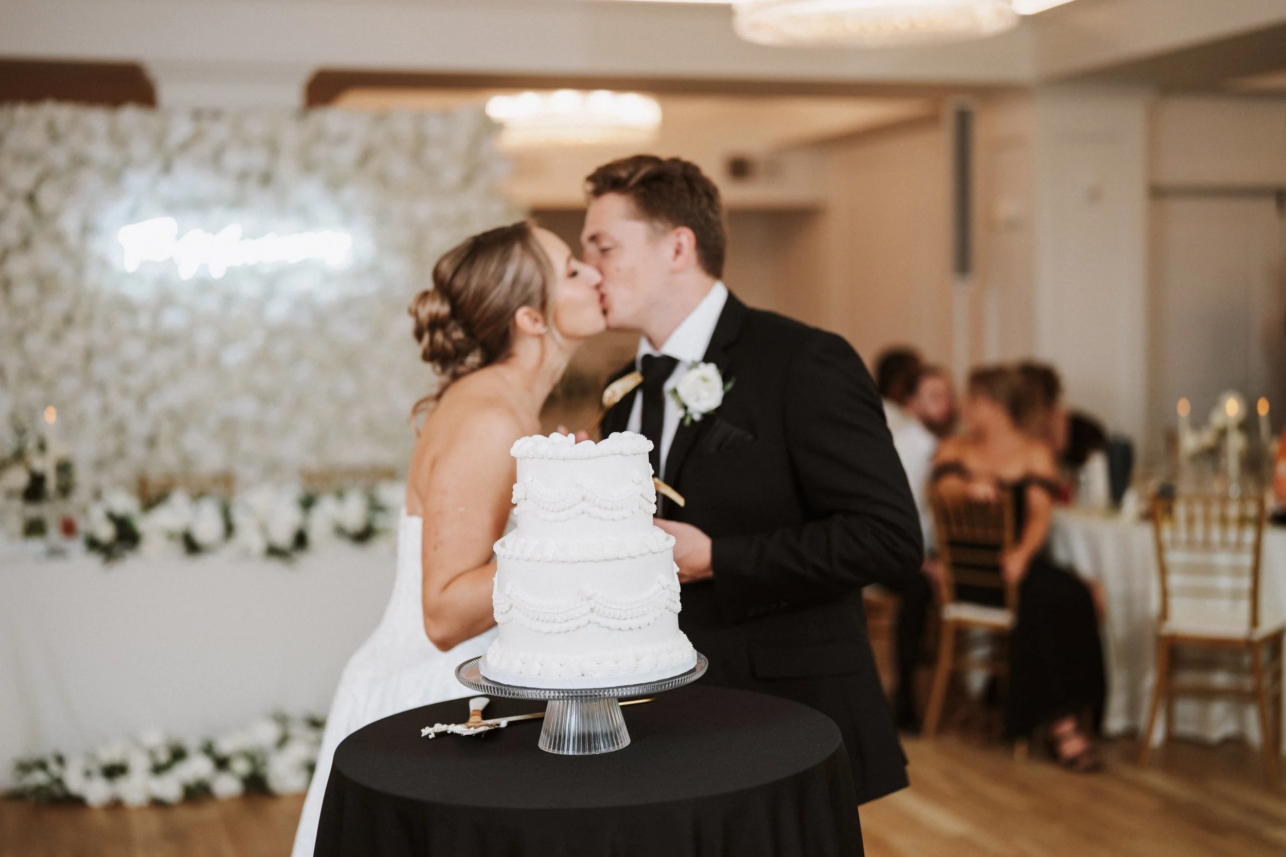 A bride and groom kiss during their wedding reception, with a wedding cake in the foreground on a black table, and guests seated at tables in the background.