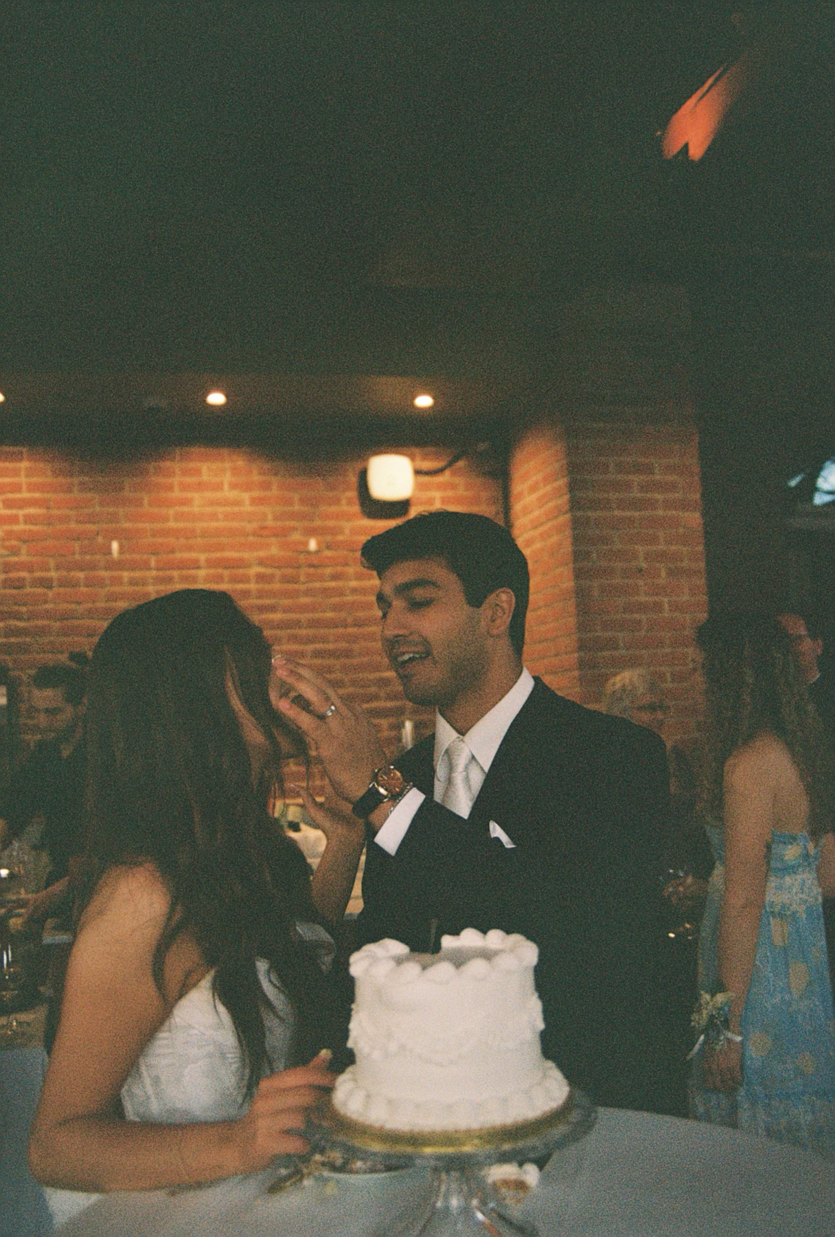 A couple celebrates at a wedding reception, sharing a joyful moment with a wedding cake in the foreground. The man is in a dark suit and the woman in a white dress, both smiling and engaging with each other in a lively indoor setting with brick walls