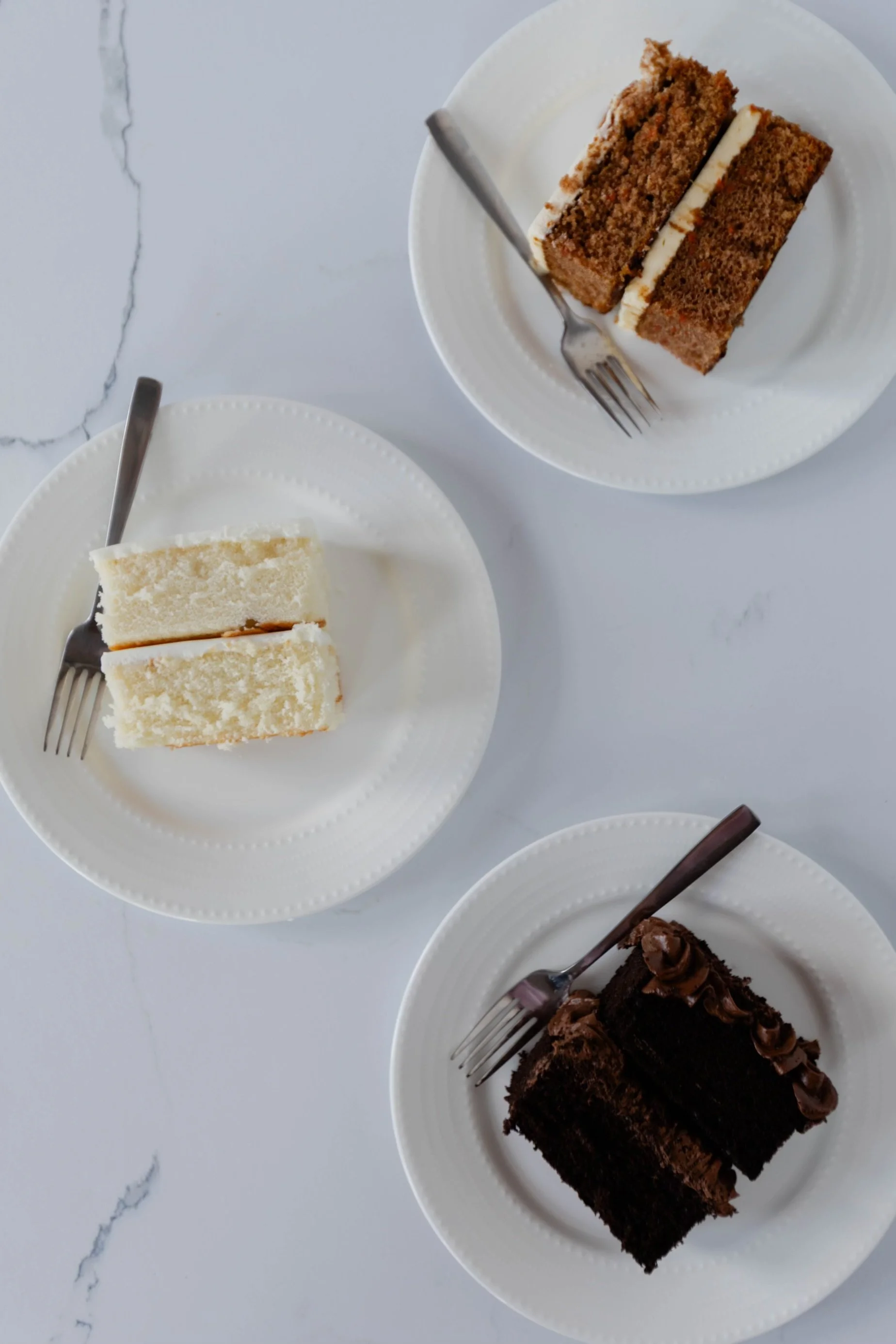 Three plates with slices of different cake types, each with a fork on white plates on a white marble surface.