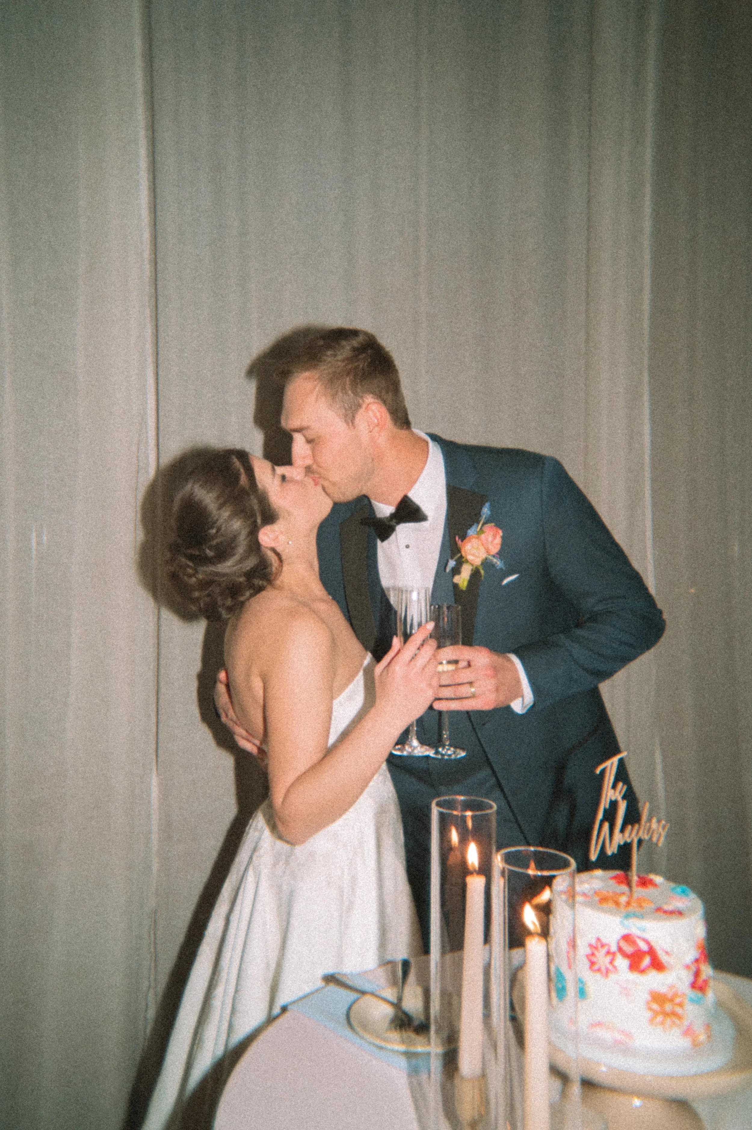A bride and groom sharing a kiss at their wedding reception, holding champagne glasses, with a wedding cake and candles on the table.