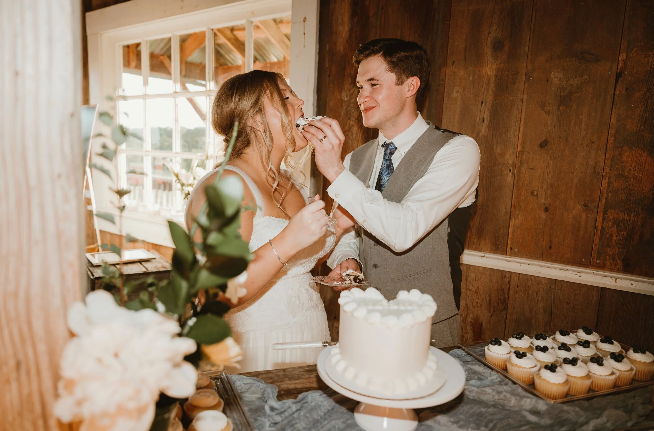 Bride and groom sharing cake at wedding reception, bride is feeding cake to groom, with wedding cake and cupcakes on table in front, rustic wooden wall background.