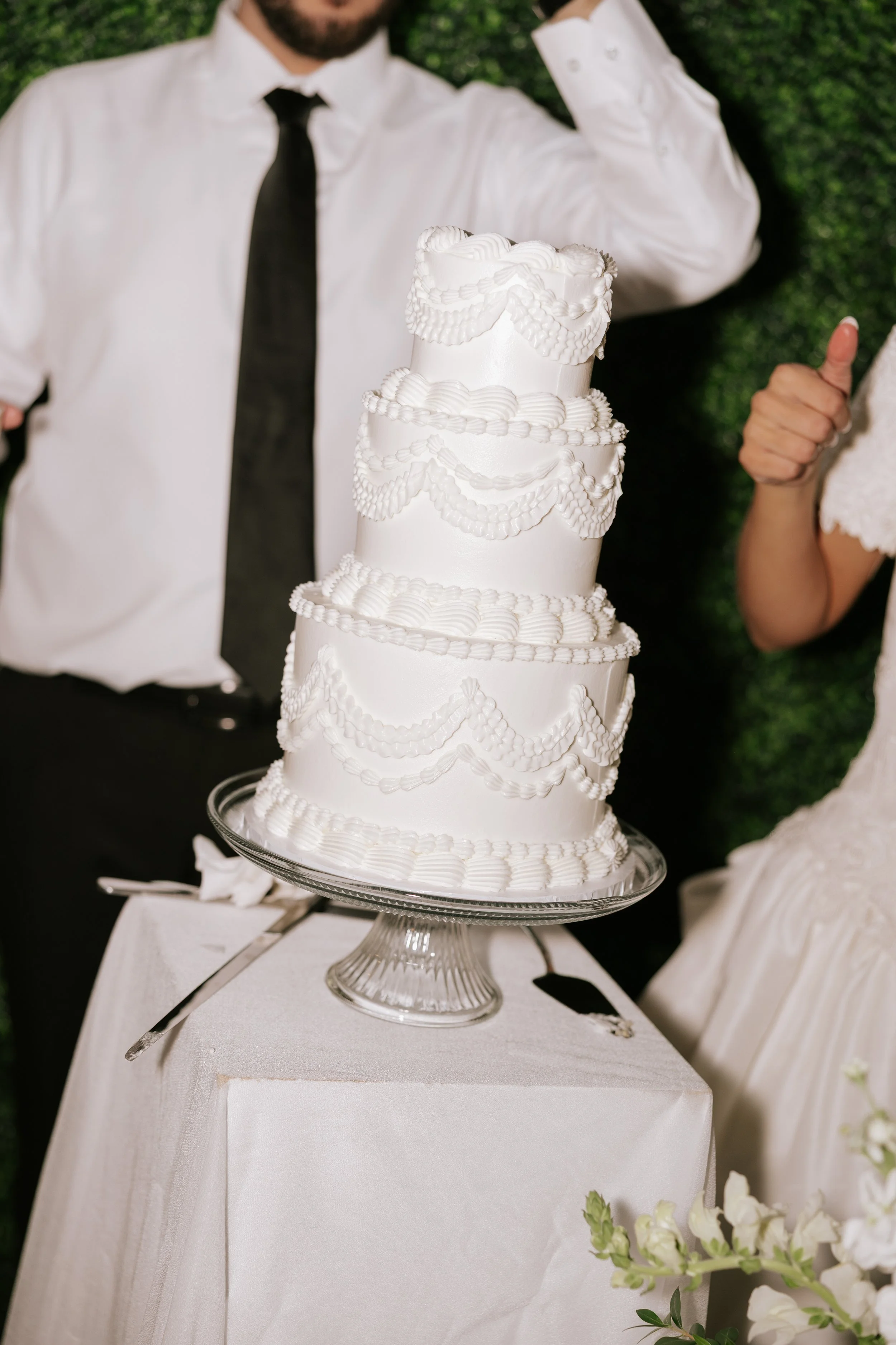 A wedding cake with five tiers decorated with white icing and piped designs, placed on a glass cake stand on a table covered with a white cloth. Partially visible are a man in a white shirt and black tie, and a woman in a white dress giving a thumbs-