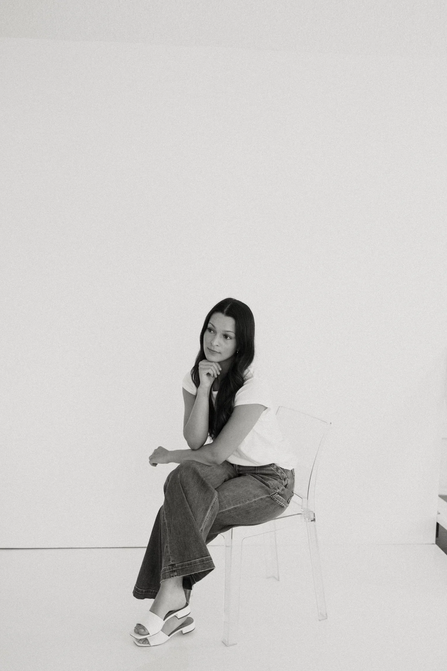 A woman with long dark hair sits on a transparent chair against a plain light background, resting her chin on her hand, dressed in a white shirt, jeans, and white sandals.