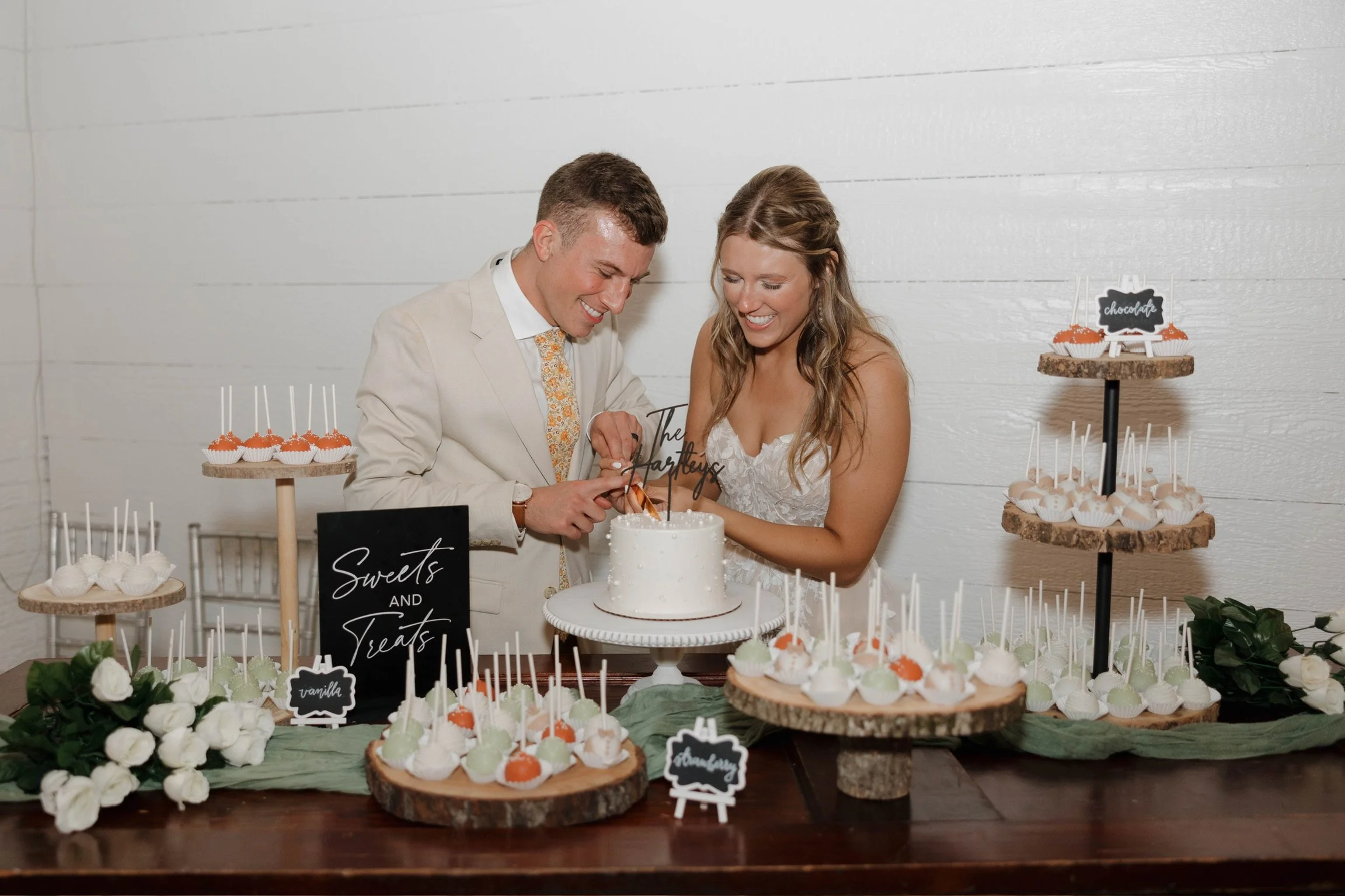 Couple cutting a cake at a wedding reception with a dessert table of cupcakes and cake pops decorated with white and orange, greenery, and a black and white sign that says 'Sweets and Treats'.