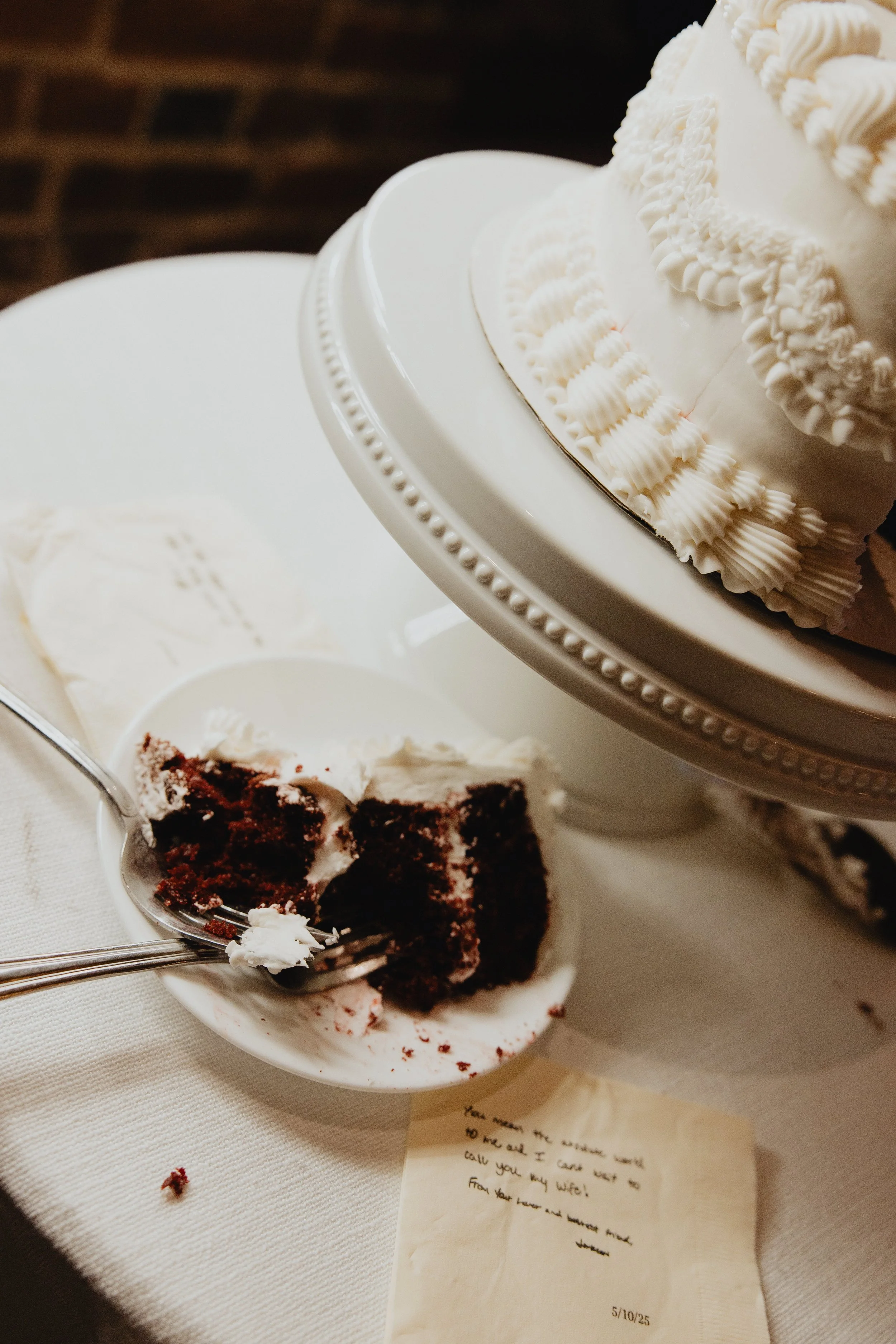 A slice of chocolate cake with white frosting on a spoon, a tiered white frosted cake on a cake stand, and a handwritten note on a napkin.