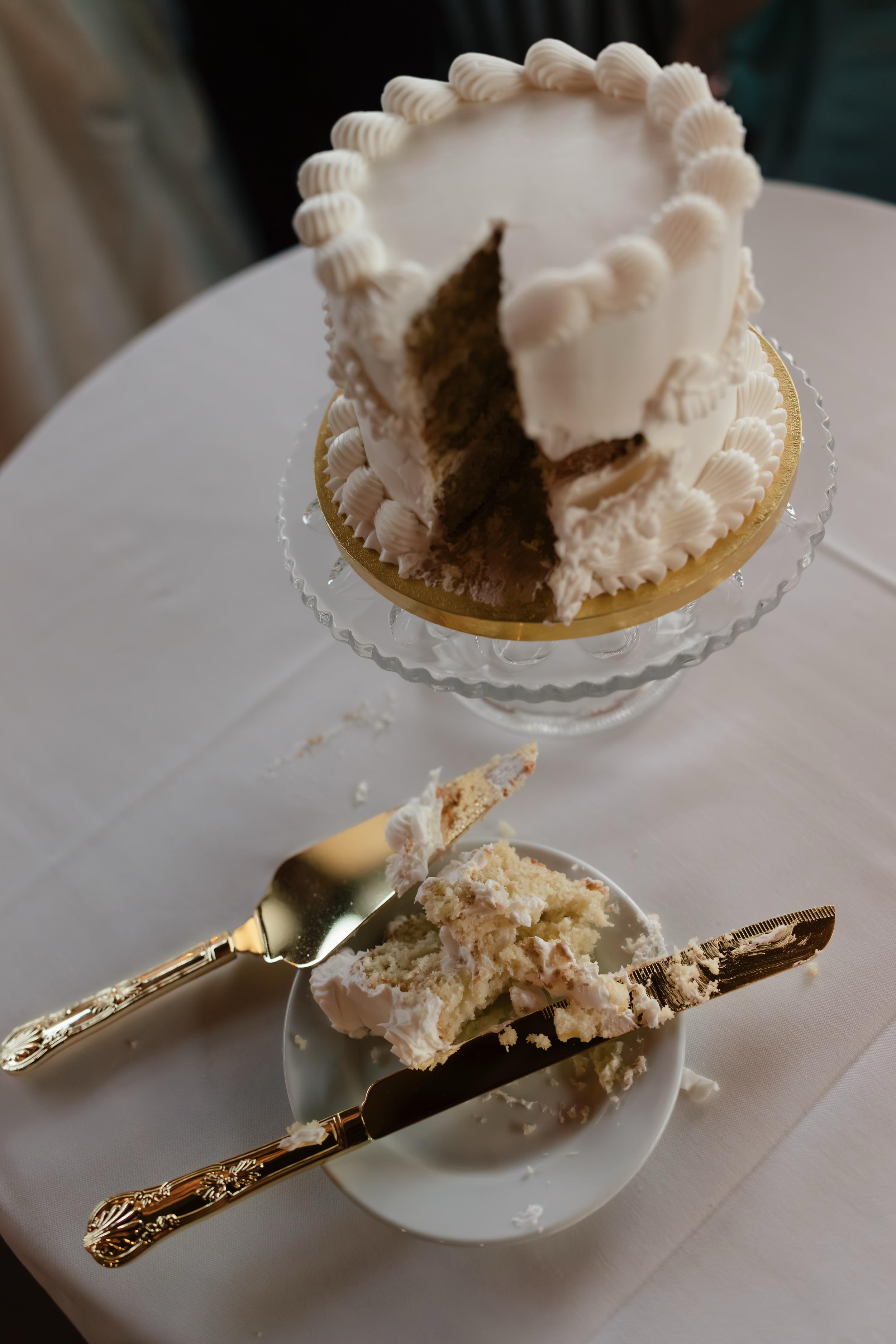 A partially eaten layered white cake with piped frosting on top, displayed on a glass cake stand; a slice has been served on a small plate with a fork, with cake crumbs and frosting remaining on the plate.