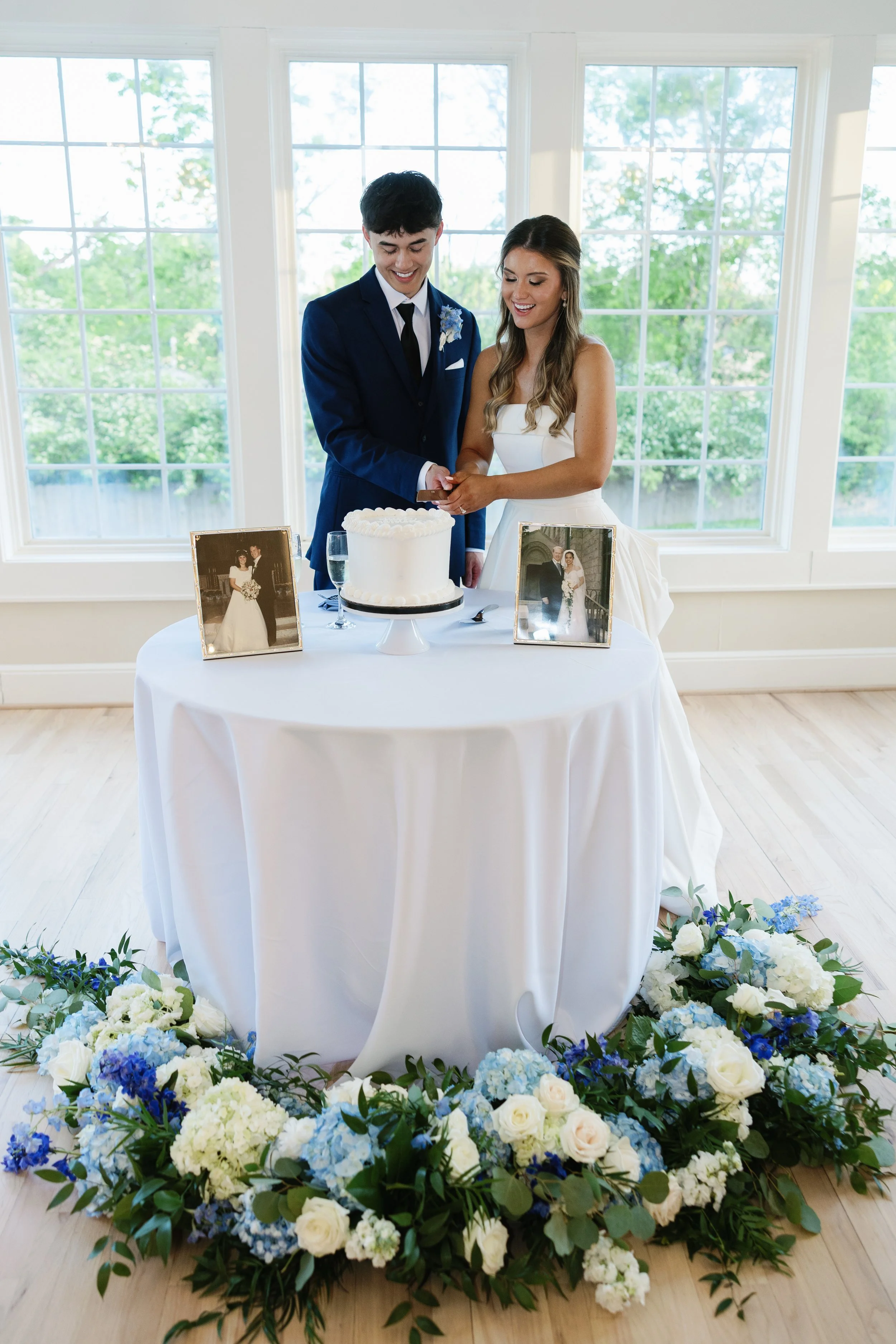 A couple in wedding attire cutting a wedding cake at reception, surrounded by floral arrangements, photos, and large windows in the background.