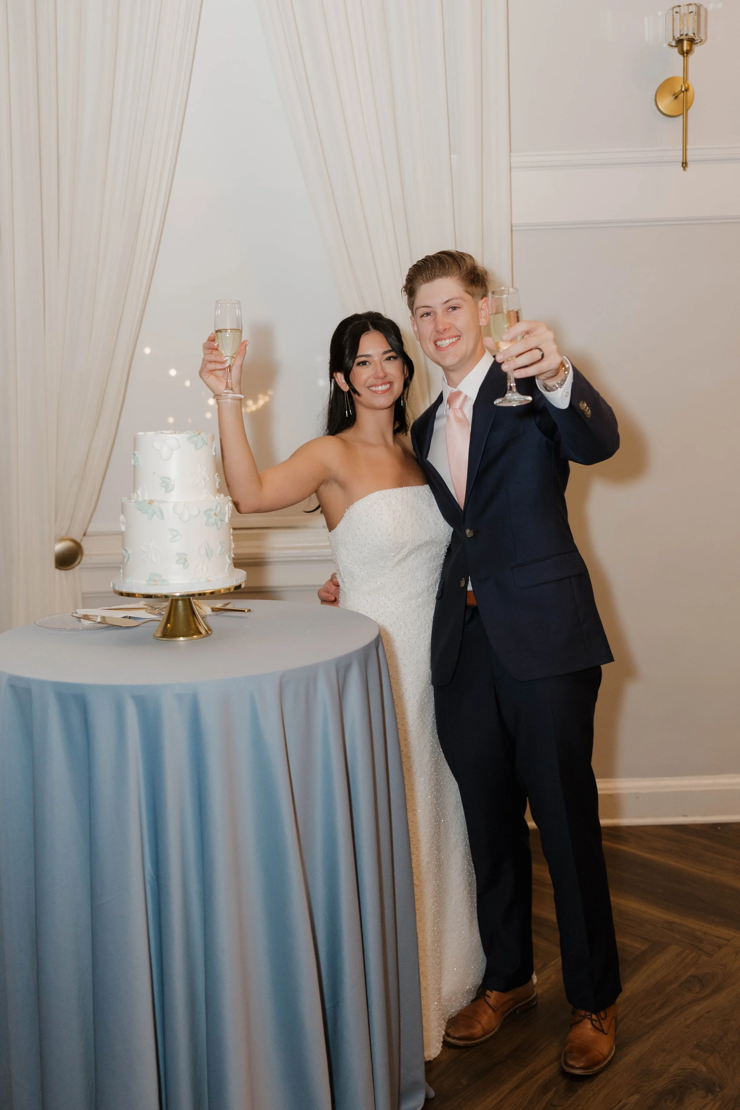 Bride and groom smiling and raising champagne glasses at their wedding reception, standing next to a tiered wedding cake on a table with a light blue tablecloth.