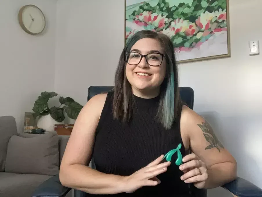 A woman with dark hair and glasses sitting in a chair, smiling, holding an orange eye mask, with a tattoo on her arm, in a living room with a wall clock, a sofa, a plant, and a colorful floral painting.