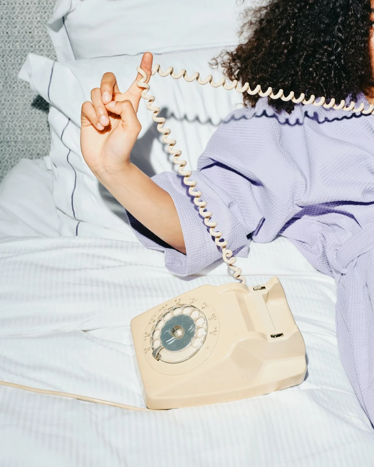 Person lying in bed, holding a vintage rotary phone's receiver with one hand, the phone resting on the bed, wearing a light purple shirt.