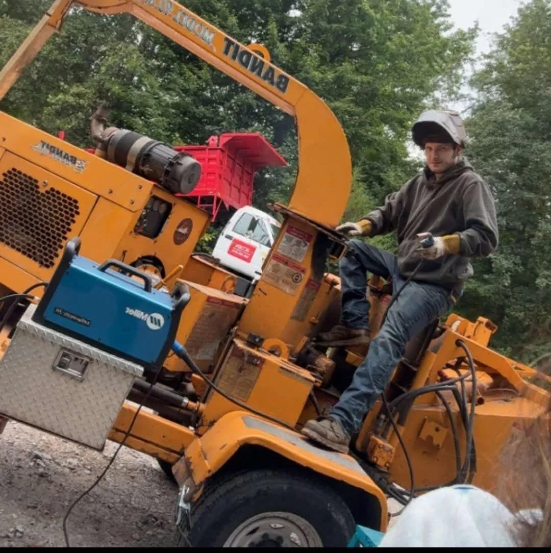 A construction worker operating a large orange wood or asphalt saw on a road during daytime, with trees and a white service vehicle in the background.