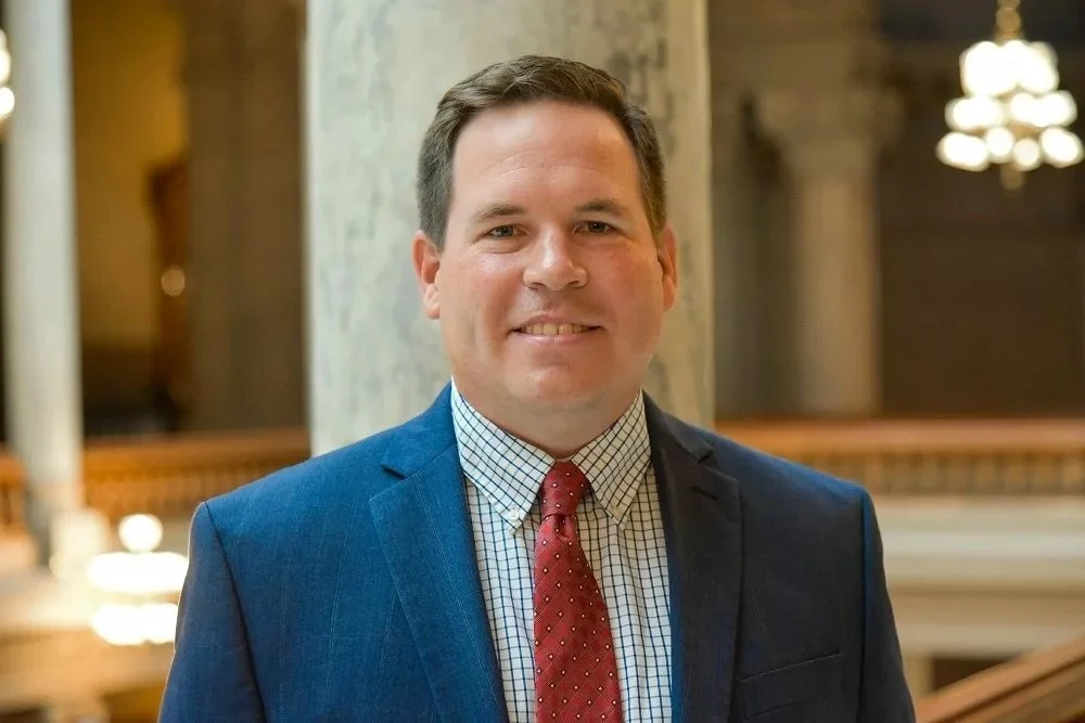 A portrait of a man in a blue suit and red tie, smiling, inside a building with ornate decor.