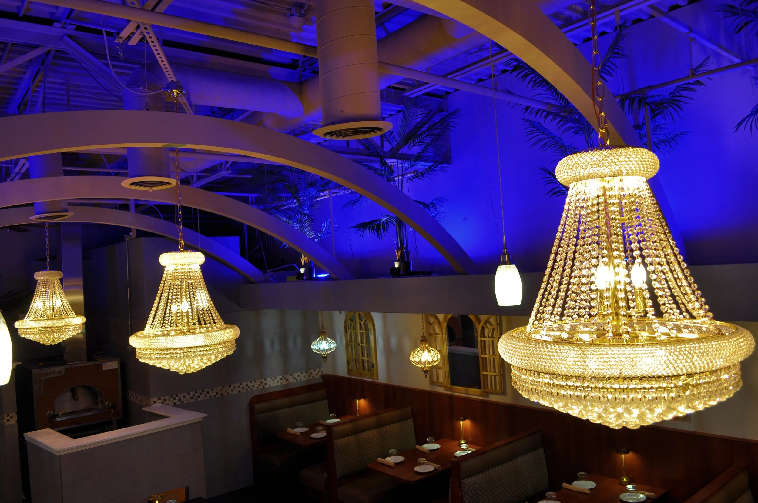 Interior of a restaurant with multiple ornate crystal chandeliers hanging from the ceiling, dim lighting, and a blue-lit ceiling with decorative plants.