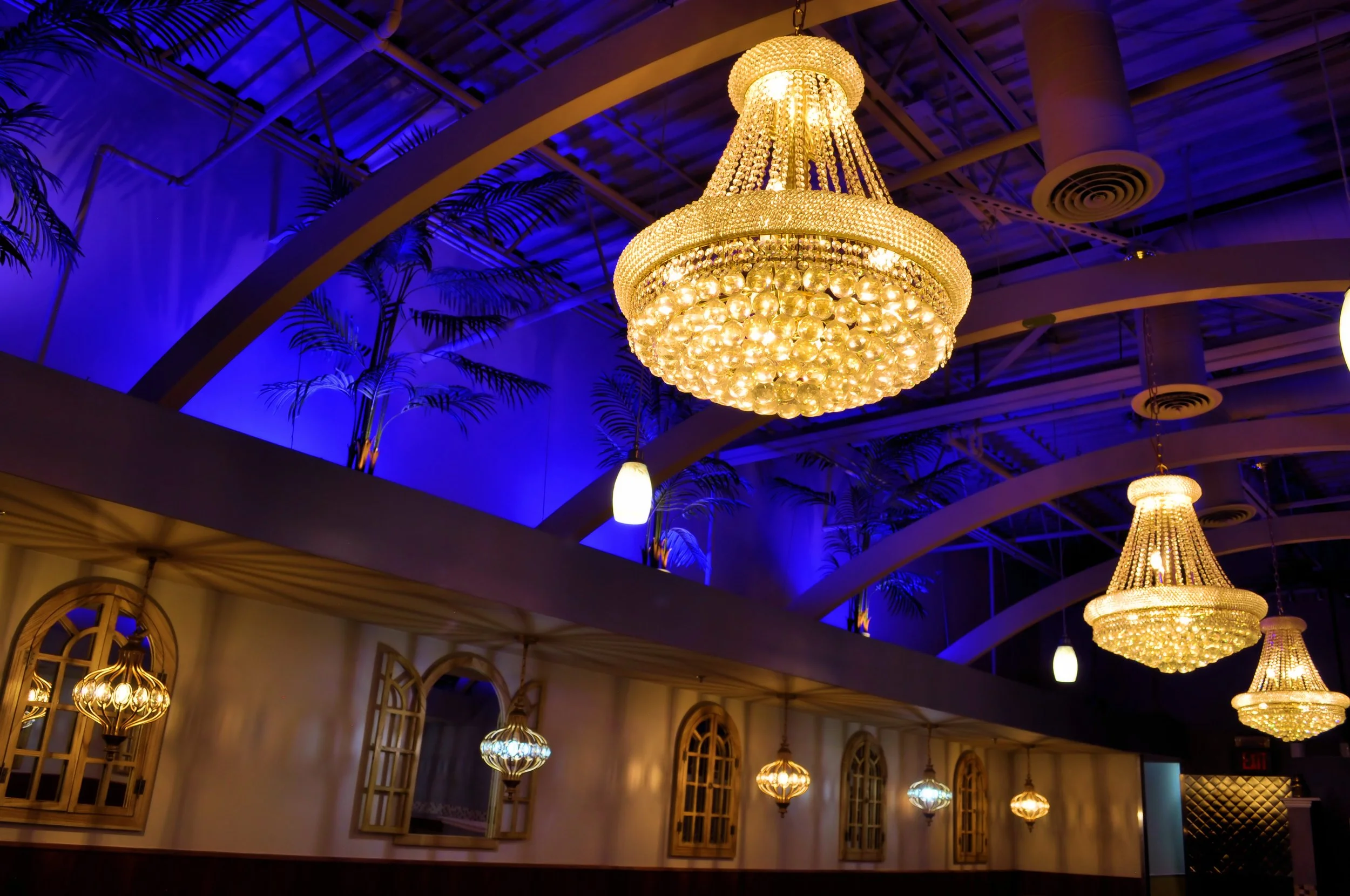 Elegant interior with ornate chandeliers hanging from a high ceiling with blue lighting and decorative palm tree art