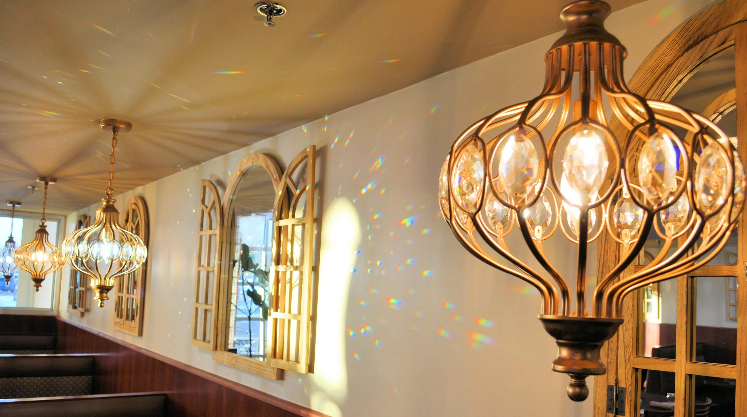 A row of ornate, gold-colored chandeliers with crystal accents hanging from a ceiling in a restaurant or café. Sunlight streams through arched windows, casting rainbow-colored reflections on the wall.