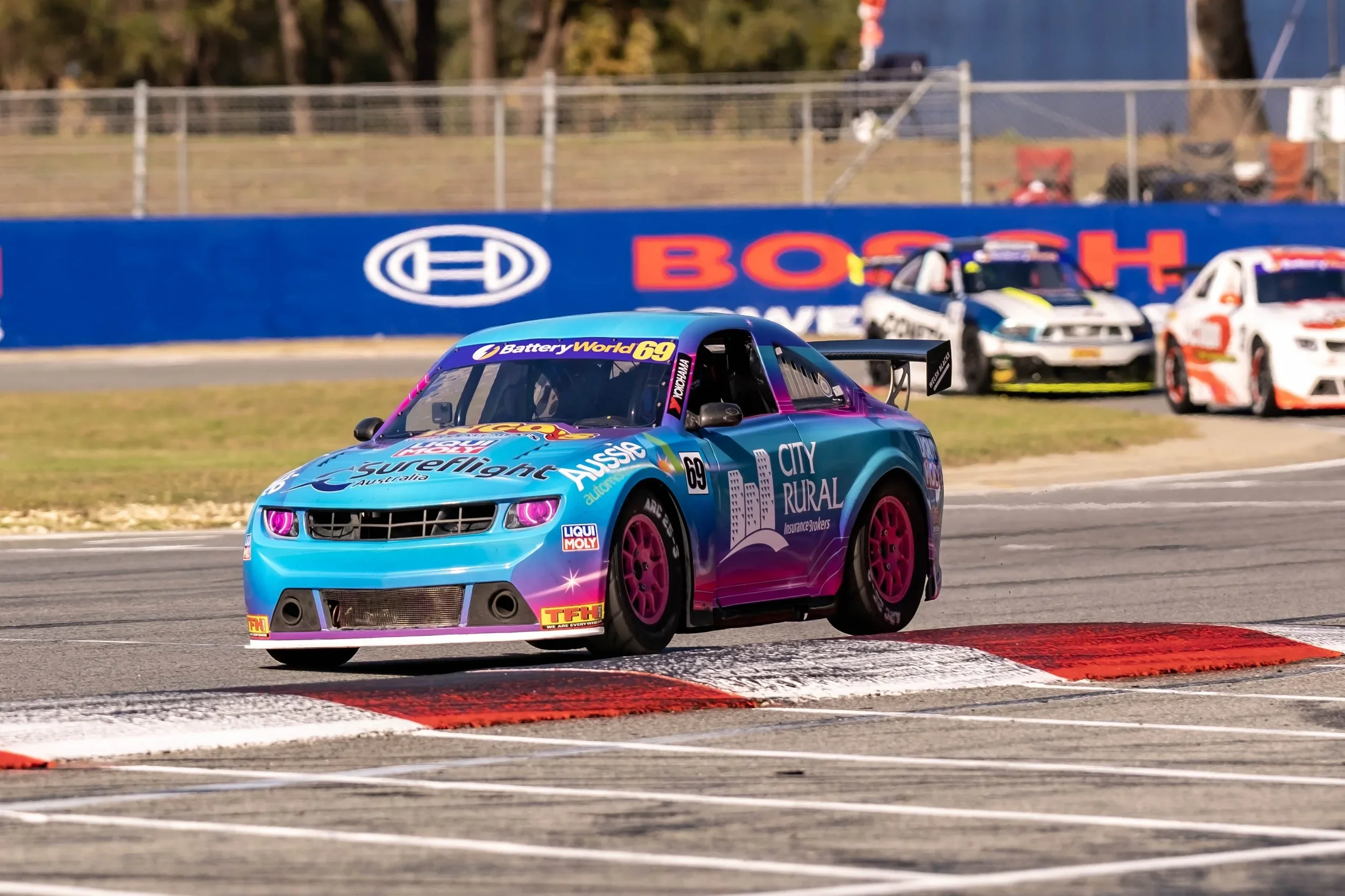 A colorful race car with pink wheels and playful sponsorship decals on a racetrack, with other race cars behind it, during a race event.