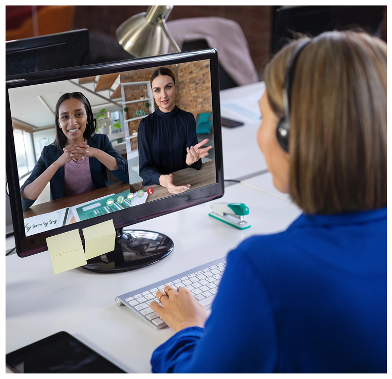 A woman in a blue blazer and headset participates in a video call with two women, one with black hair and the other with blonde hair, in an office setting.