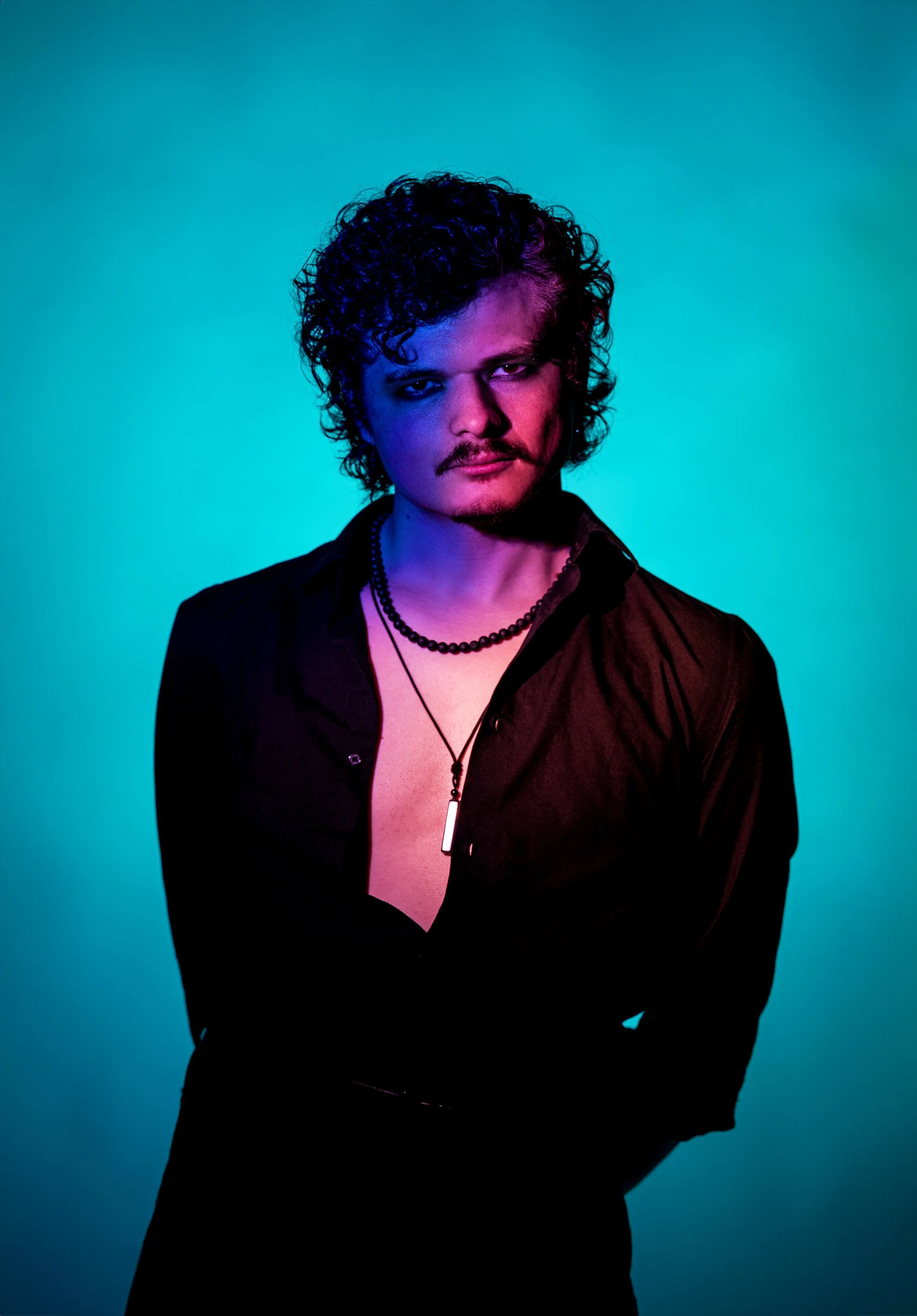 A man with curly dark hair and facial hair, wearing an unbuttoned black shirt, multiple necklaces, and standing against a brightly lit, gradient background of blue and purple hues.