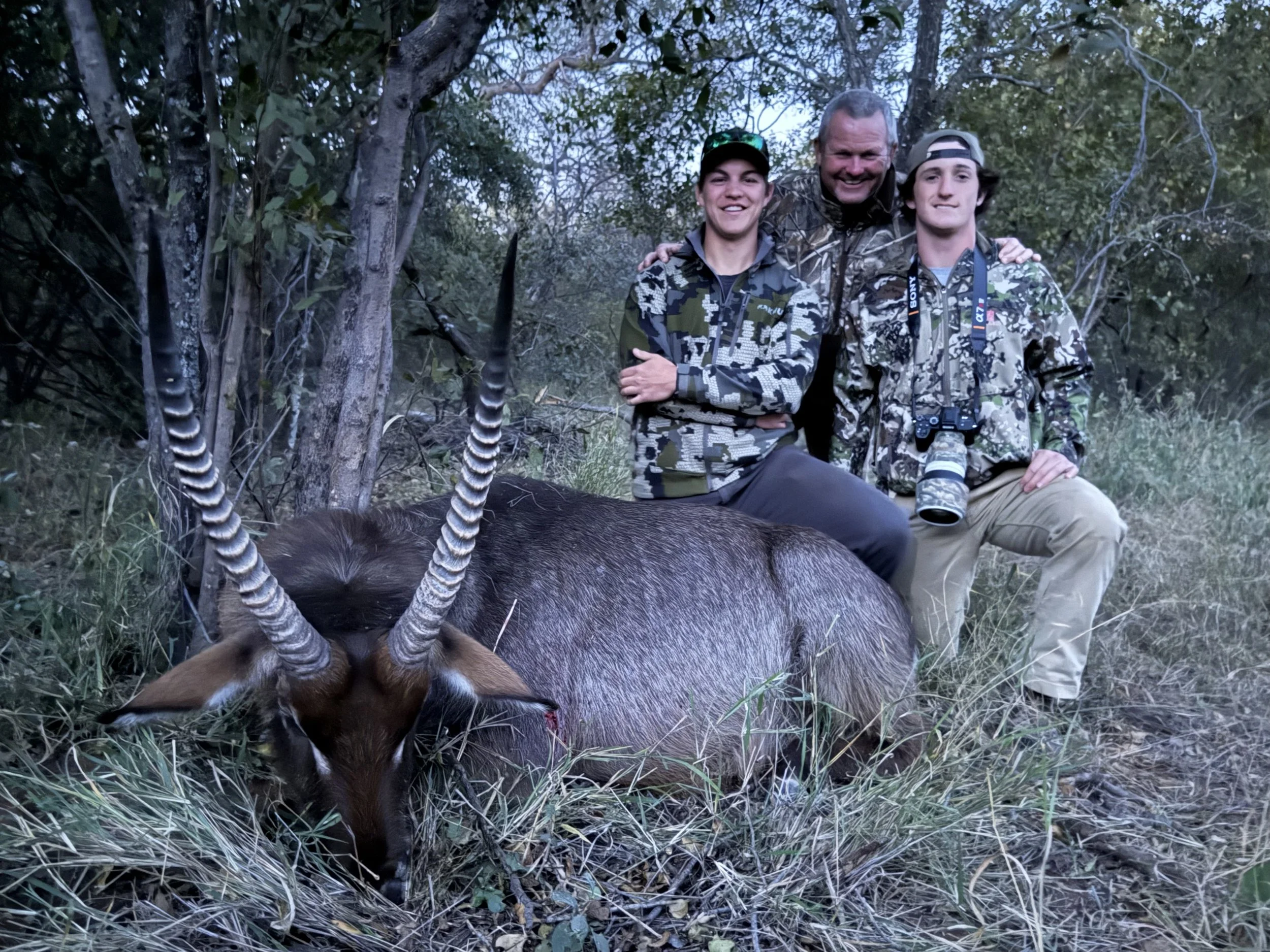 Three people in camouflage clothing kneeling behind a dead deer with antlers, outdoors in a wooded area.