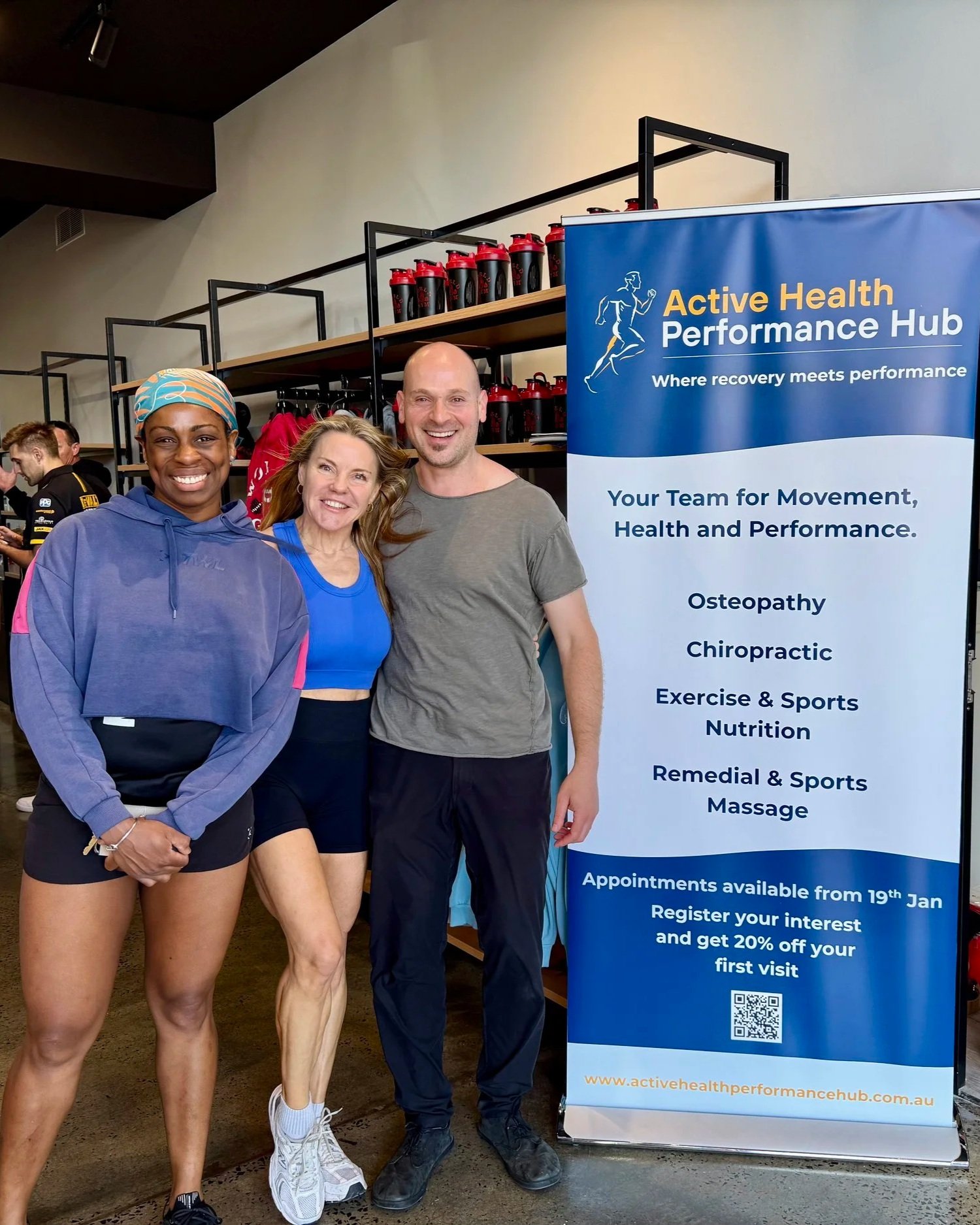 Three people smiling and standing together indoors next to a blue banner for Active Health Performance Hub, which offers services like osteopathy, chiropractic, exercise & sports nutrition, and sports massage. Active Health Performance Hub Bentleigh