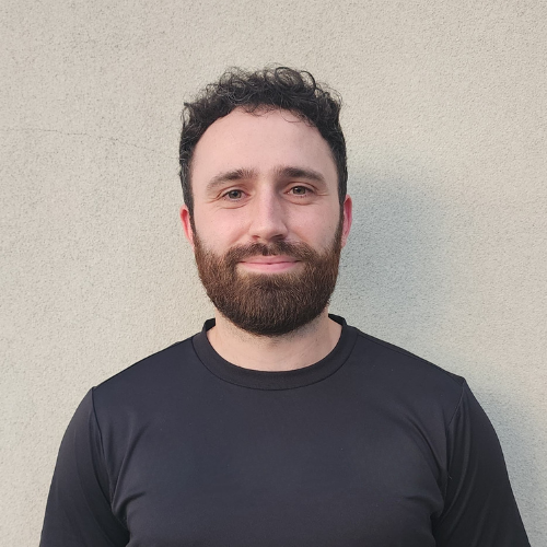 Liam Stanely Remedial and Sports Massage - Portrait of a man with curly dark hair and a beard, wearing a black shirt, standing against a plain light-colored wall. Active Health Performance Hub Bentleigh East