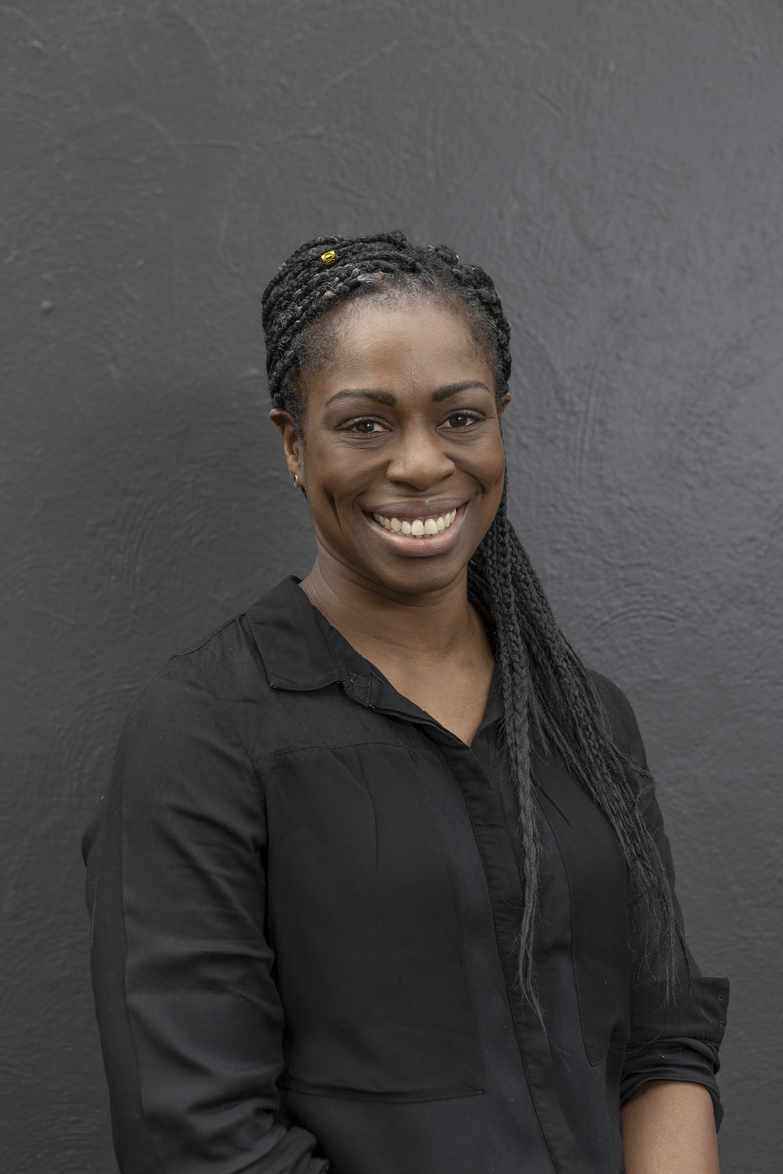 Stella Borland Remedial Massage Therapist Portrait of a smiling Black woman with braided hair, wearing a black shirt, standing against a dark gray textured background. Active Health Performance Hub Bentleigh East