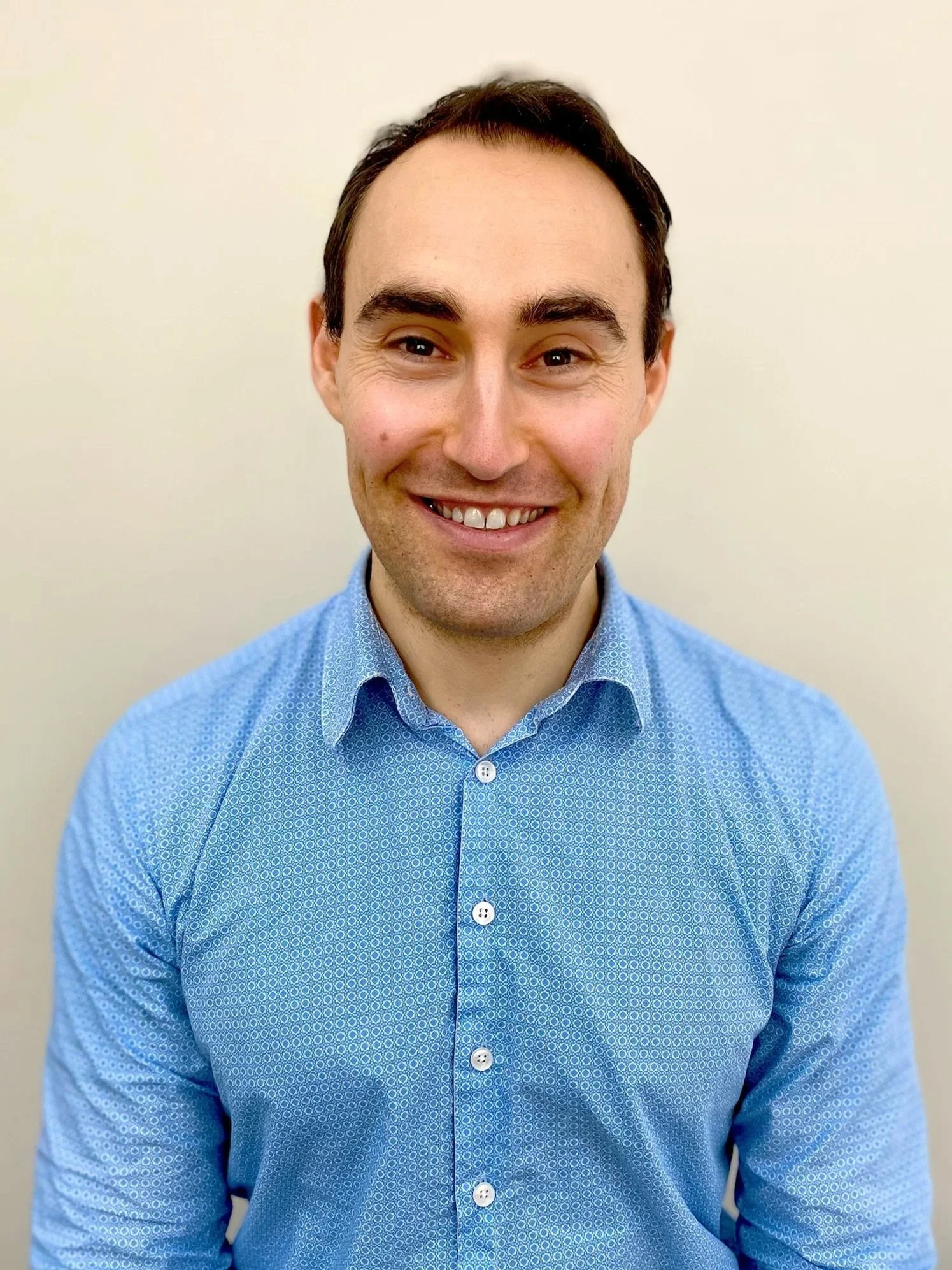 Sports Osteopath Dr David Cork. A smiling man with light skin and dark hair, wearing a light blue, patterned button-up shirt, standing against a plain light-colored background. Active Health Performance Hub Bentleigh East