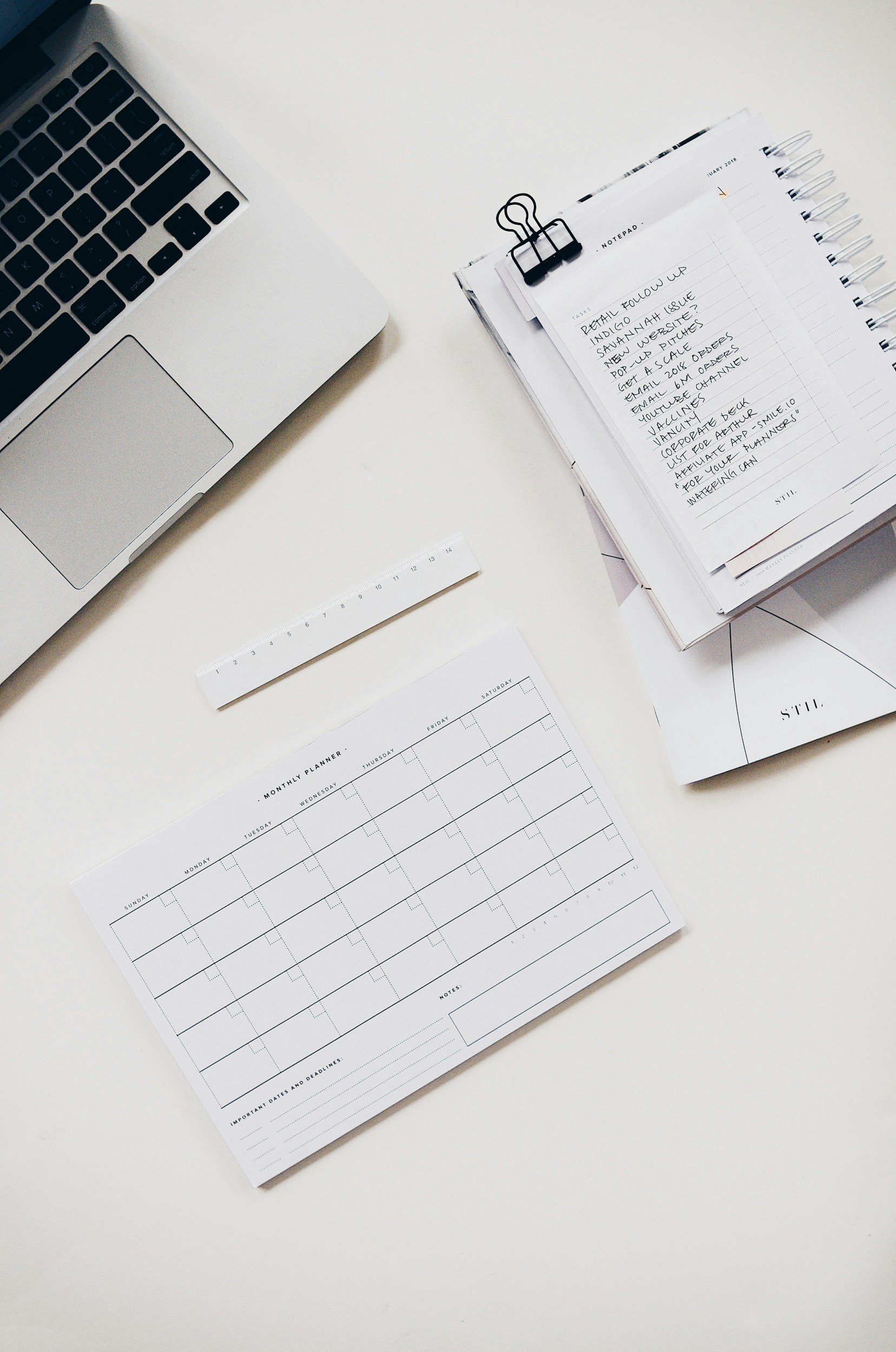 A white desk with a silver laptop, a stack of papers clipped with a black binder clip, a printed weekly calendar, and a white ruler.