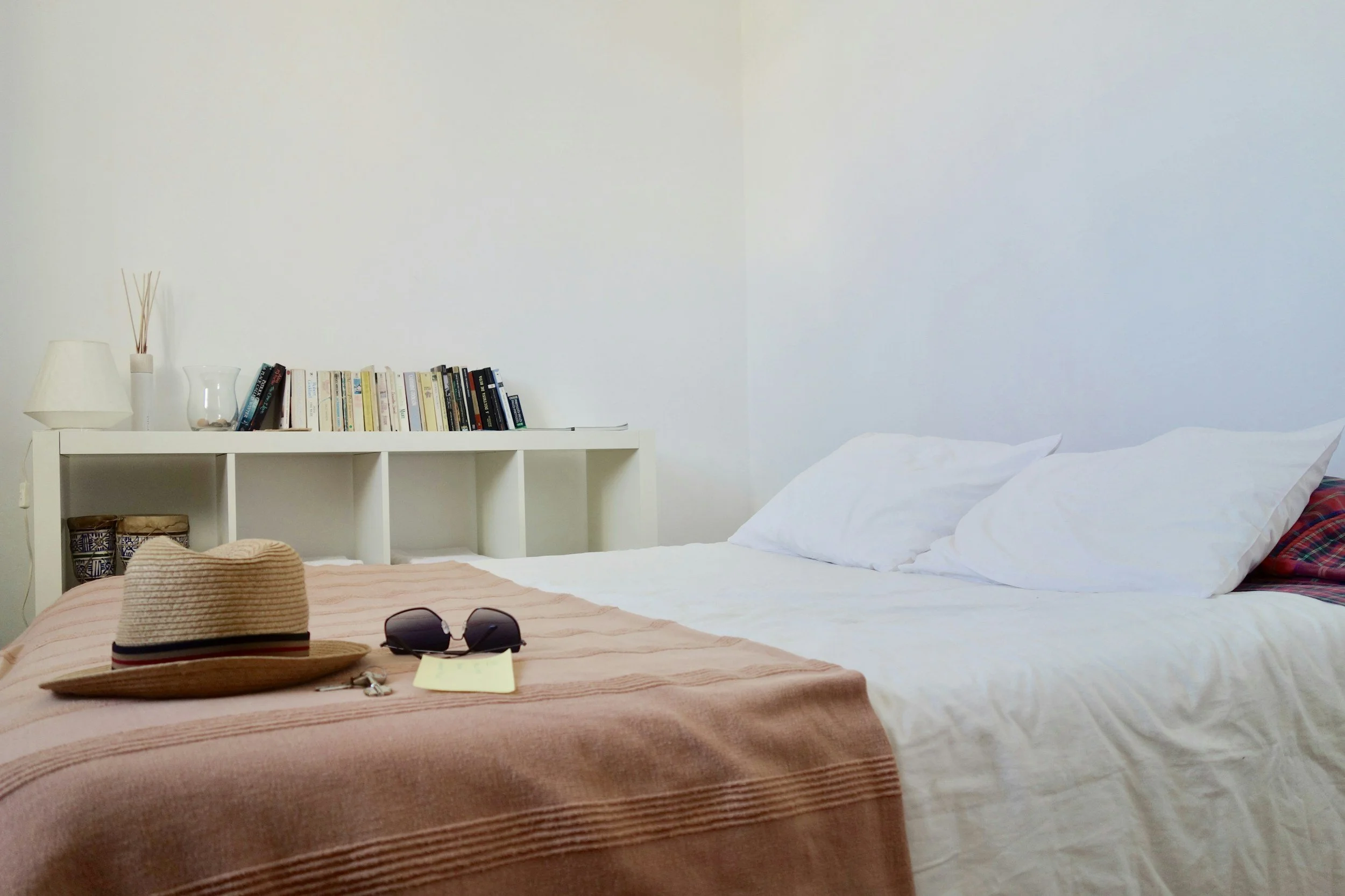 A neatly made bed with white sheets and pillows, a pink blanket, a straw hat, sunglasses, and keys on top. In the background, a white shelf with books, a lamp, and decorative items.