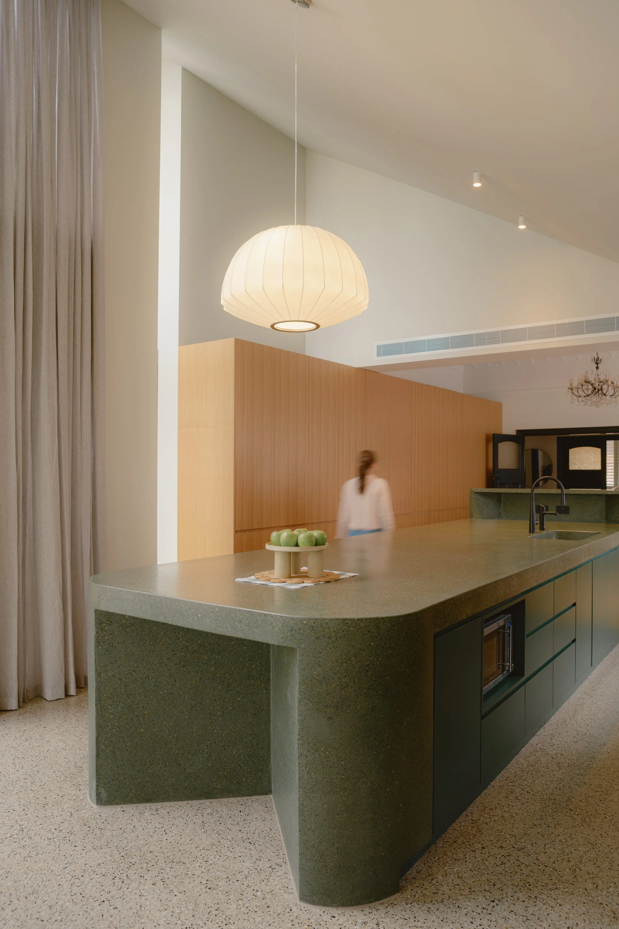 Modern kitchen with a large Mortadello terrazzo green island, hanging paper lantern light, wooden wall panel, and a person walking in the background.