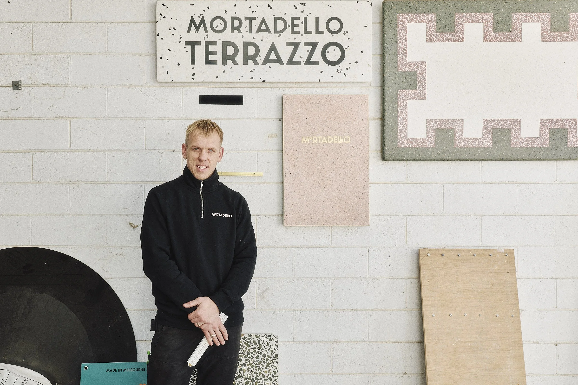 Michael Lesinskis standing in front of a white brick wall with various Mortadello Terrazzo sample panels, wearing a black jacket with the logo 'Mortadello'.
