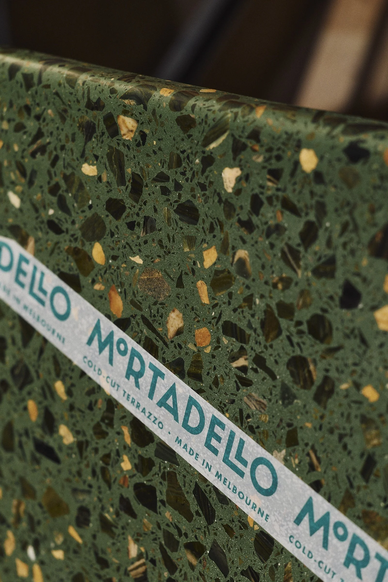 Close-up of a green terrazzo surface with multicolored stone fragments and a white strip of text reading 'MORTEDELLO' in blue letters, indicating it is a cold-cut terrazzo made in Melbourne.