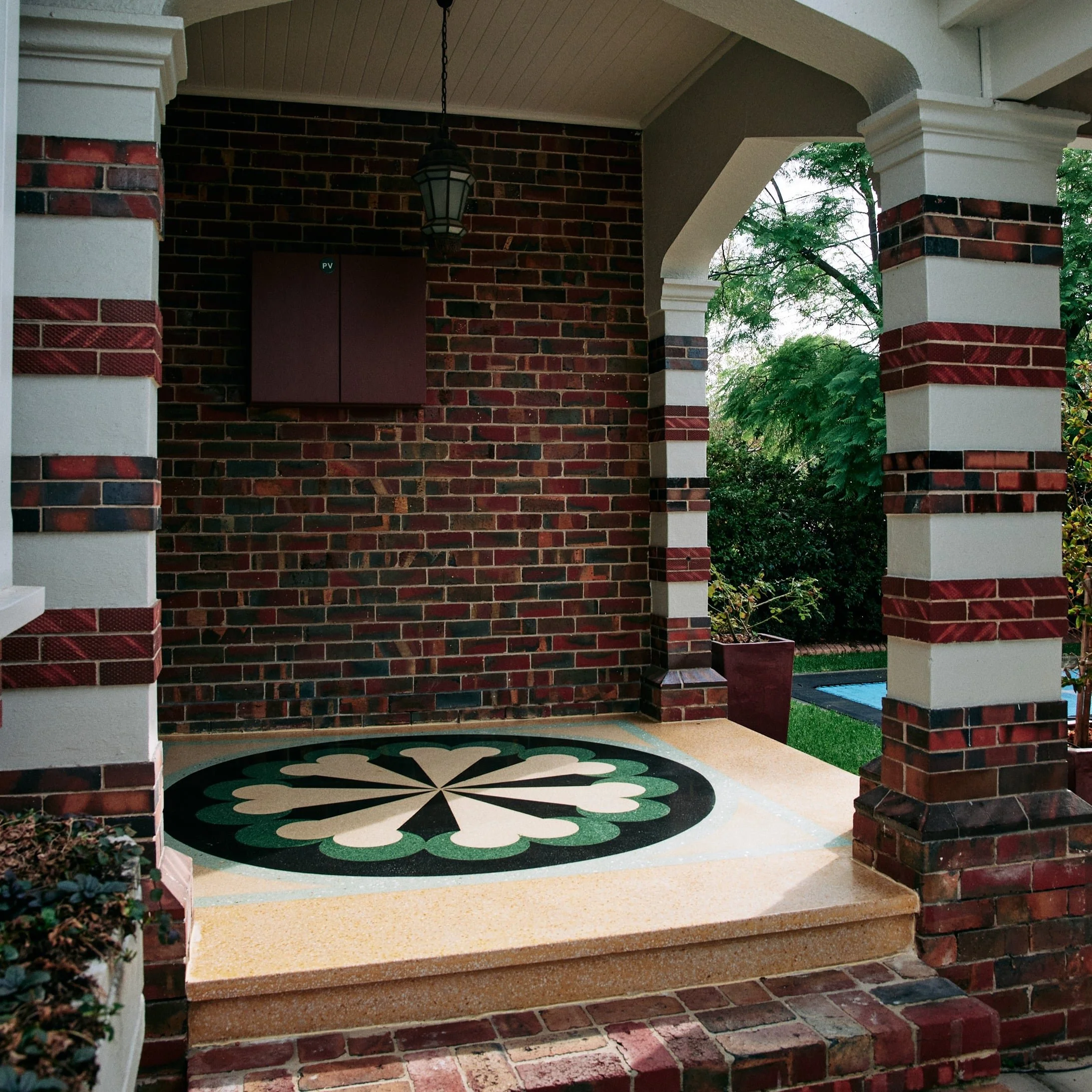 Porch with brick columns and a tile floor featuring a green and cream floral terrazzo pattern, surrounded by lush greenery.