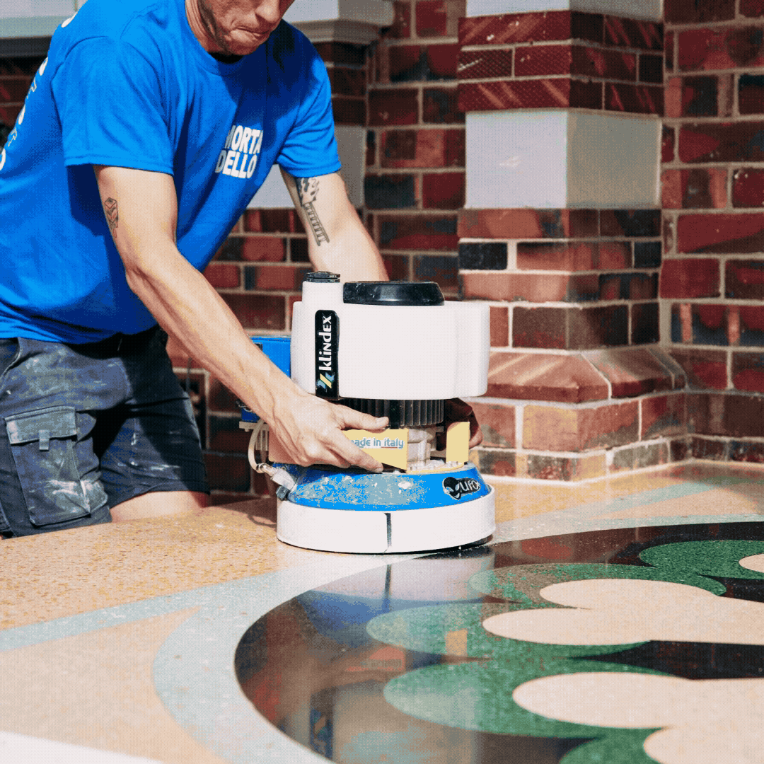 A man in a blue shirt is polishing the terrazzo porch with a machine outside near a brick wall.