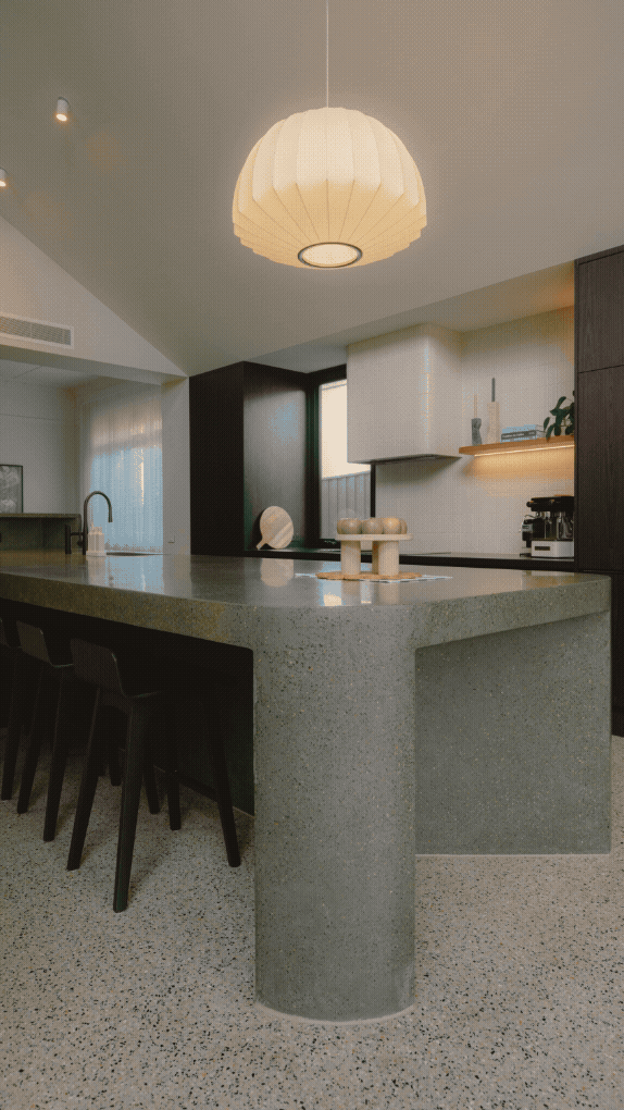 Modern kitchen with a large green terrazzo island, black bar stools, a hanging cream-colored pendant light, and minimalist decor.