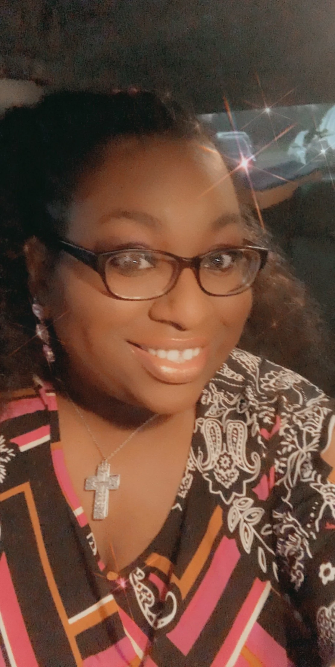 Close-up selfie of a smiling woman with glasses, earrings, and a cross necklace, inside a vehicle at night with cars visible in the background.