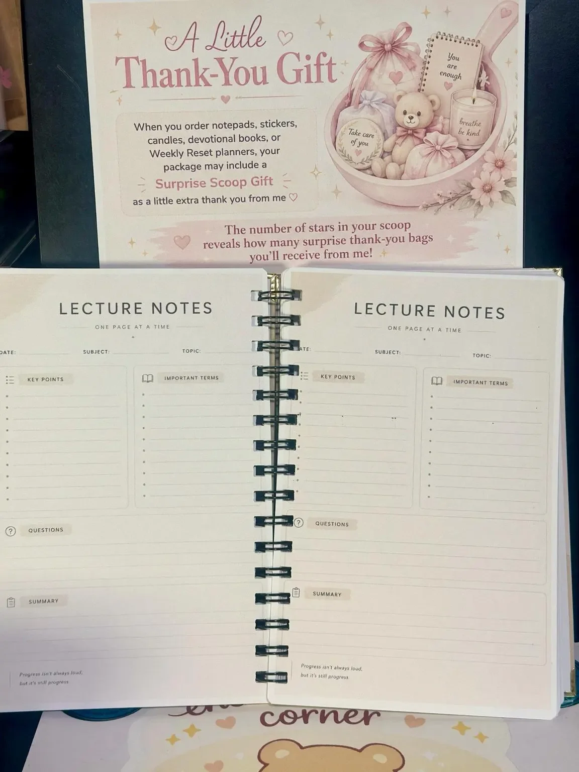 A spiral-bound notebook with two pages titled 'Lecture Notes' on each side, featuring sections for key points, important terms, questions, and a summary. Behind the notebook is a card or poster with a pink, heart-themed design that reads 'A Little Thank-You Gift', with a teddy bear, candles, and pastel-colored gift bags, indicating a theme of appreciation and surprise gifts.