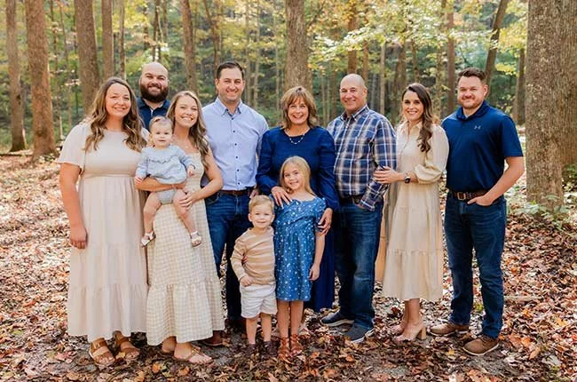 Family photo of ten people, including adults and children, standing outdoors in a wooded area with autumn leaves.