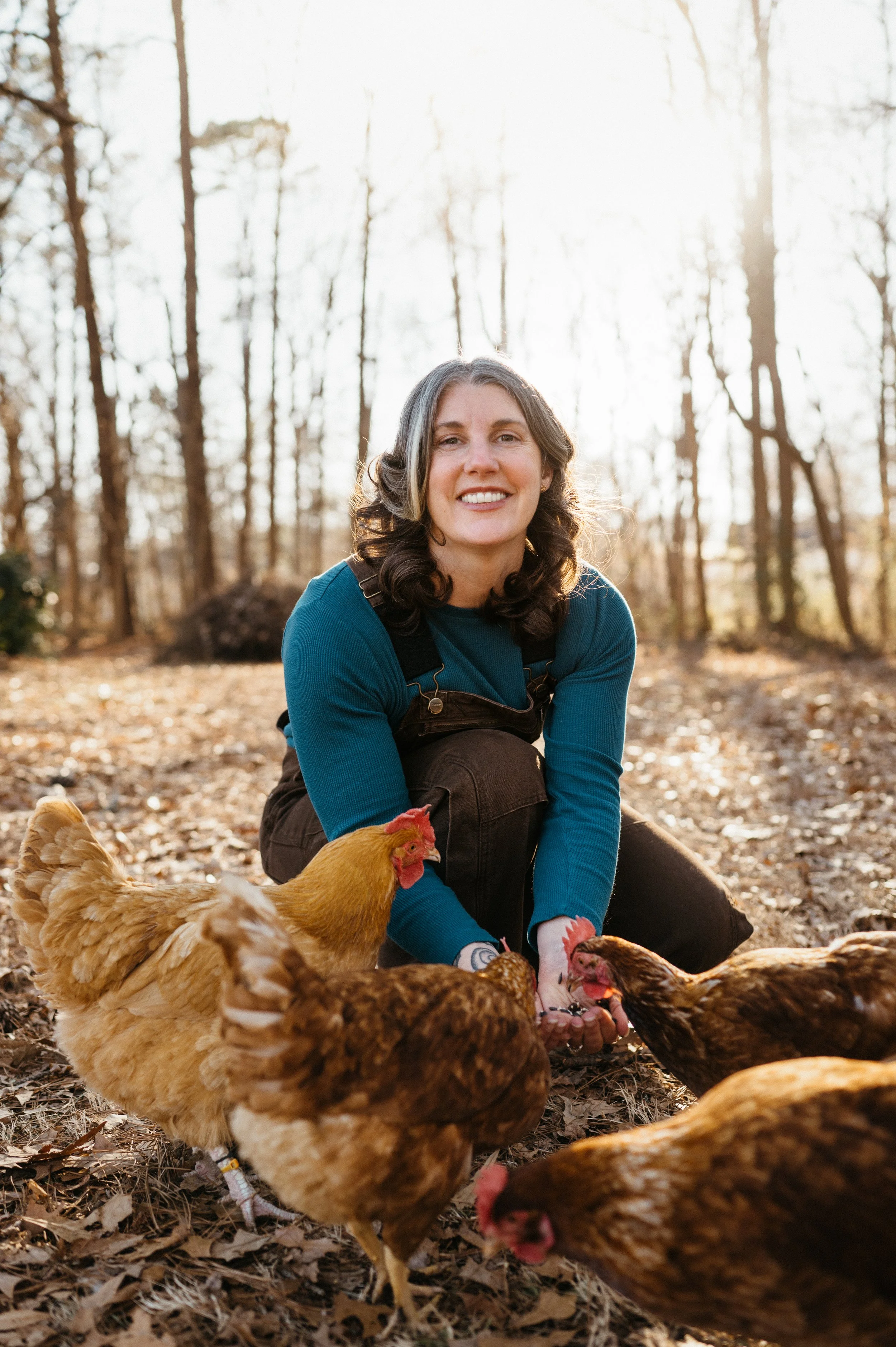 A woman smiling and crouching in a wooded outdoor area surrounded by chickens.